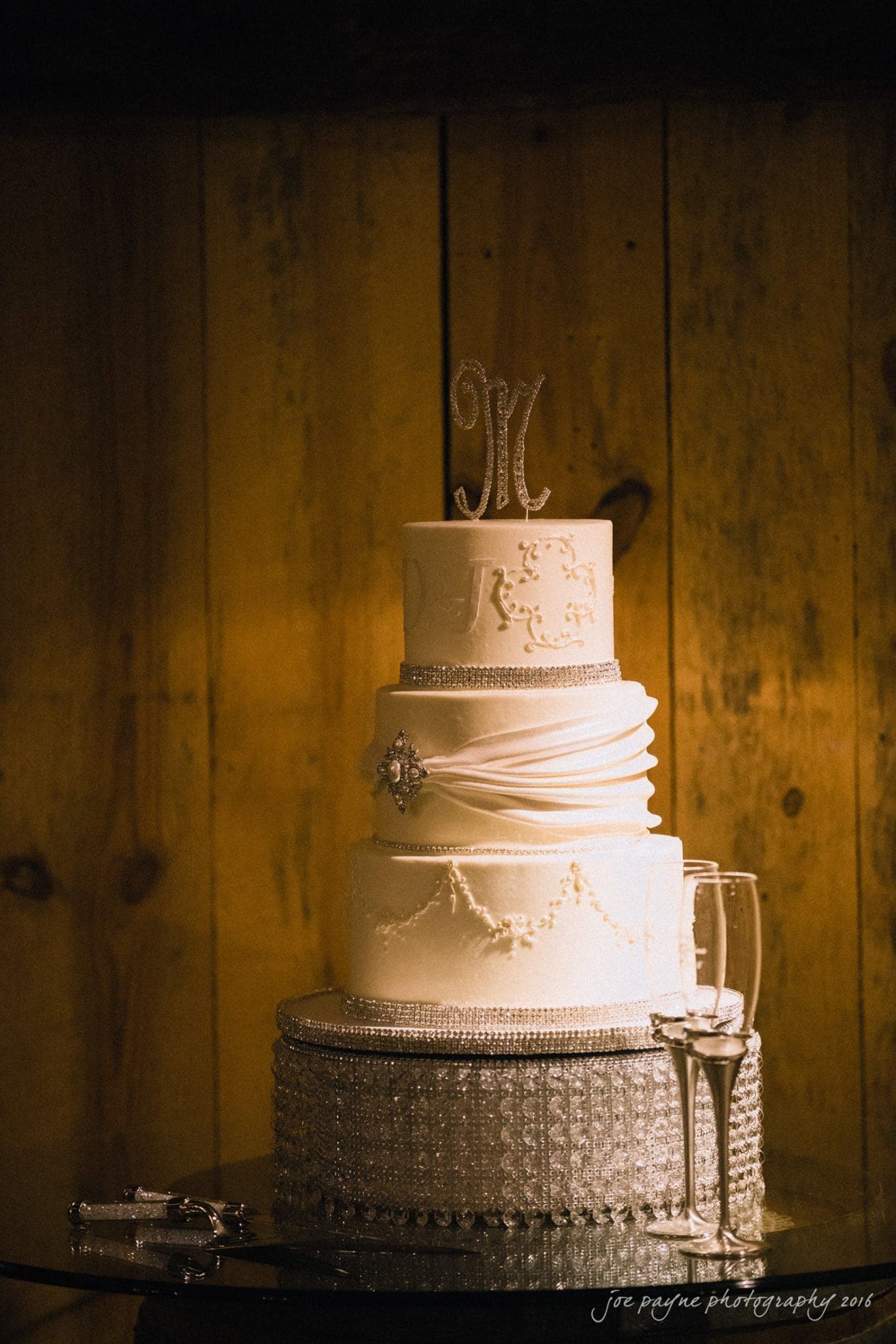 shady wagon farm wedding photography - danielle & josh A four-tier white wedding cake decorated with elegant patterns, pearls, and a monogram topper. The cake rests on a silver stand beside two champagne flutes, set against a warm, rustic wooden background.