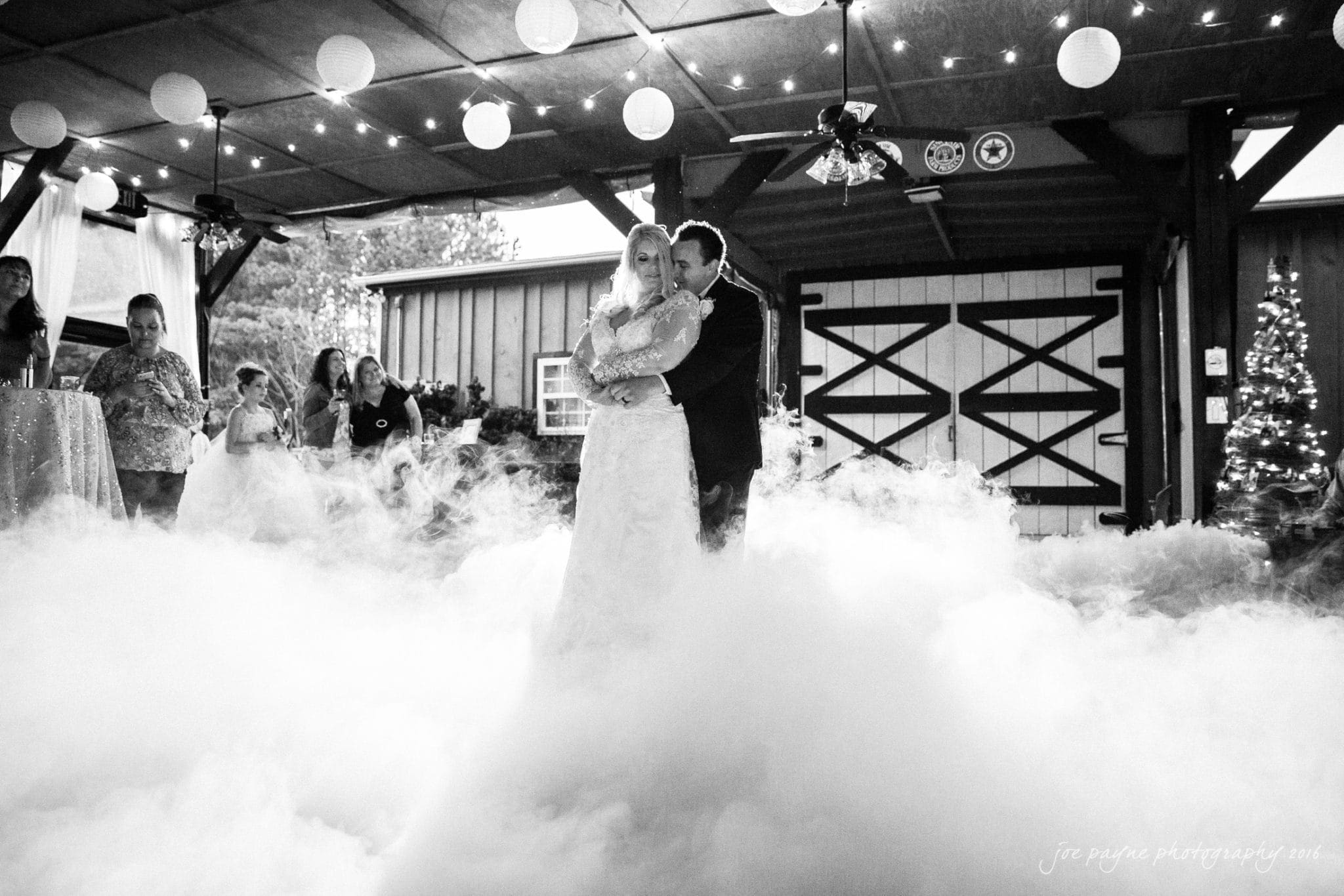 shady wagon farm wedding photography - danielle & josh A bride and groom enjoy their first dance amidst billowing mist under a rustic barn with string lights and paper lanterns. Guests watch from the background, and a decorated Christmas tree adds a festive touch.