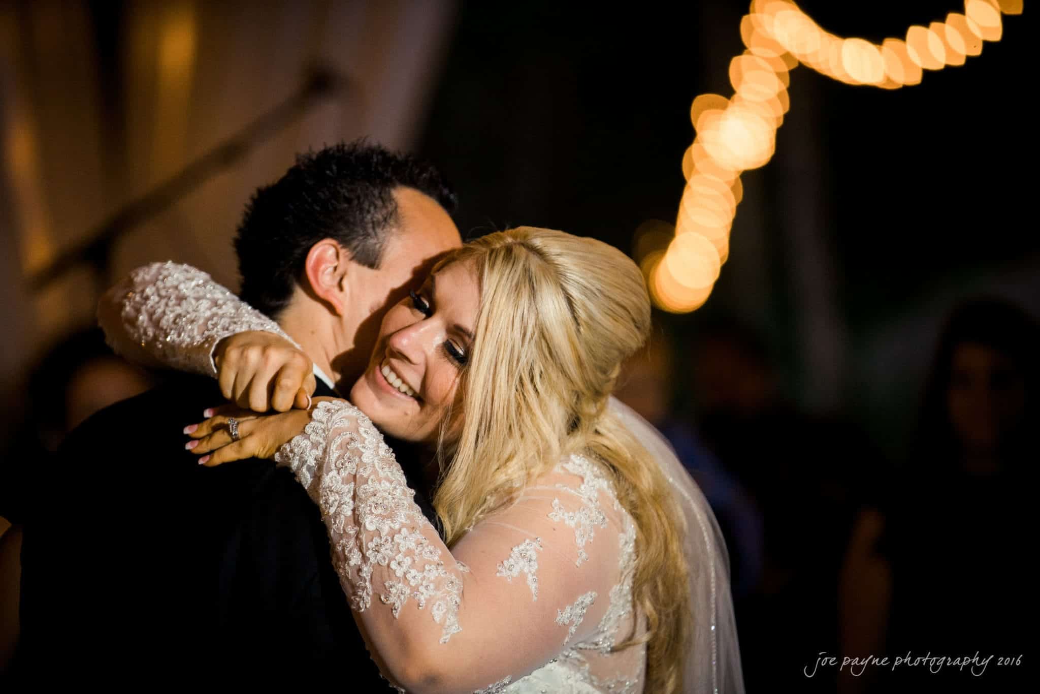 shady wagon farm wedding photography - danielle & josh A bride and groom share a joyful embrace on the dance floor. The bride is smiling with her eyes closed, wearing a lace wedding dress, and the groom is holding her close. Soft, warm lighting with bokeh effects creates a romantic atmosphere.