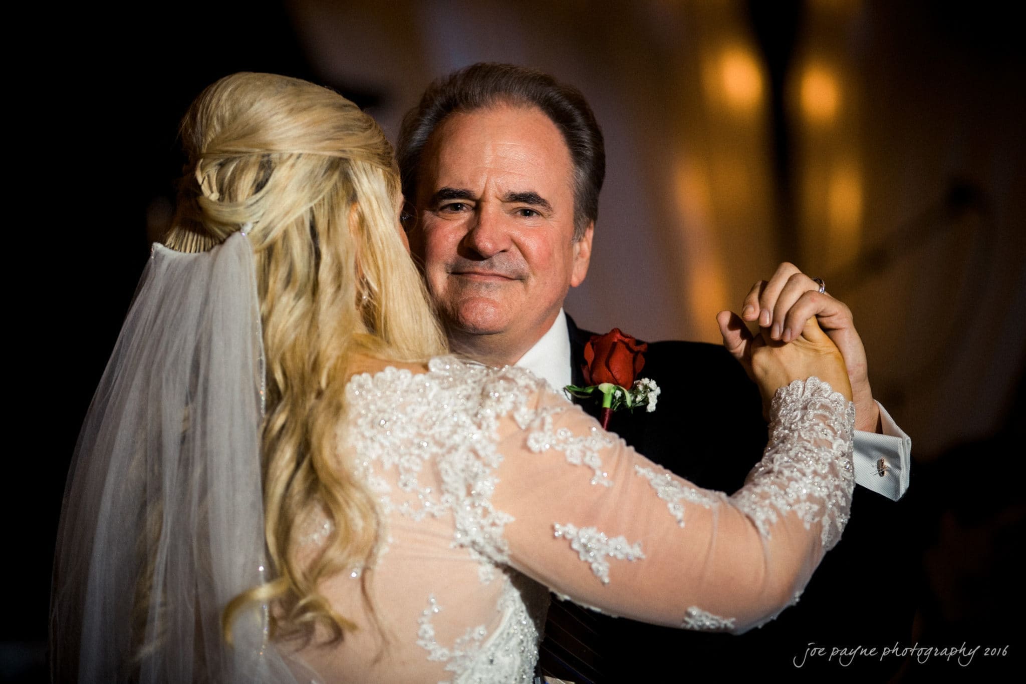 shady wagon farm wedding photography - danielle & josh A man and a woman share a dance. The woman, in a white lace dress and veil, faces away from the camera. The man, in a suit with a red boutonniere, smiles gently as they dance under soft lighting.