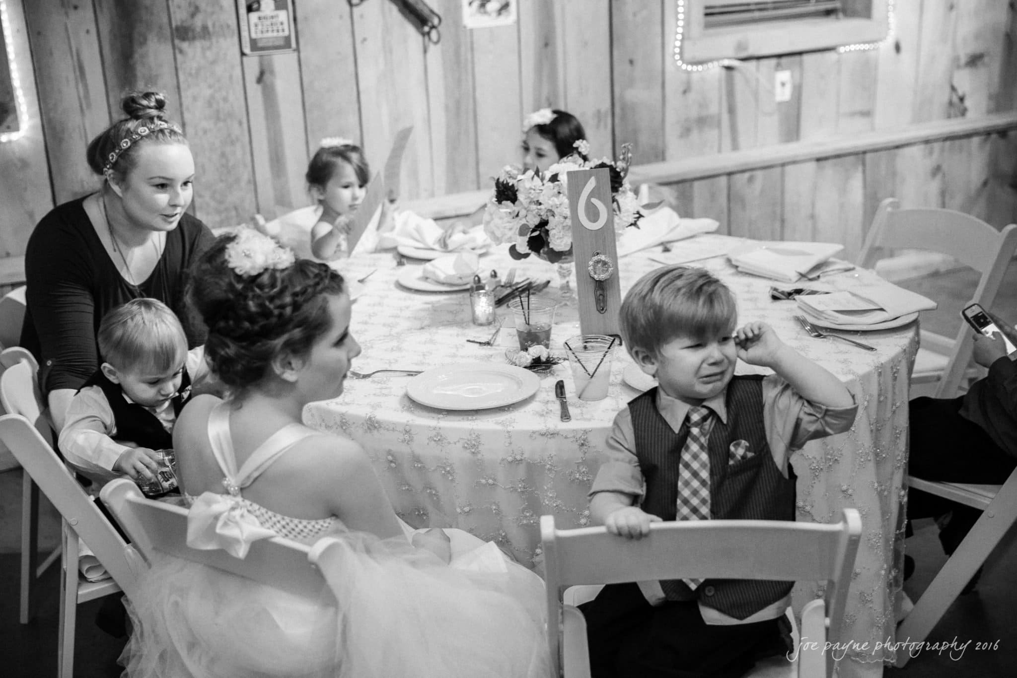 shady wagon farm wedding photography - danielle & josh A black-and-white photo of a wedding reception. Several children are seated around a table with a floral centerpiece and a table number 6. One child in a suit is visibly upset, while others look on or are focused elsewhere.
