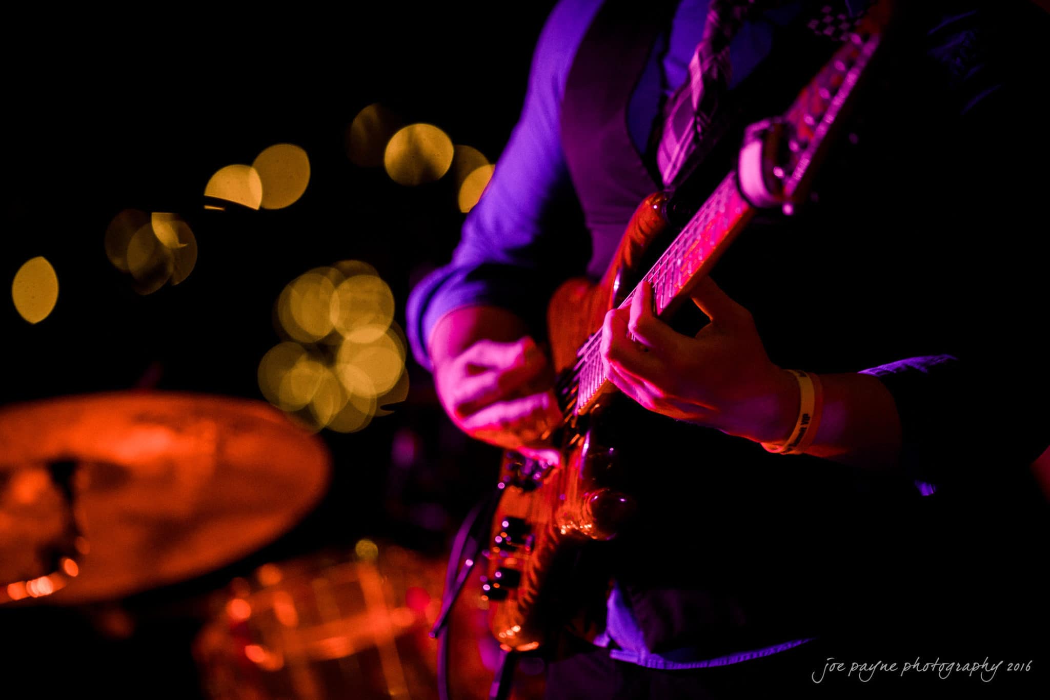shady wagon farm wedding photography - danielle & josh A musician plays an electric guitar under colorful stage lights, with a blurred background showing a drum set and glowing orbs. The atmosphere is energetic and vibrant, highlighting the performances intensity.