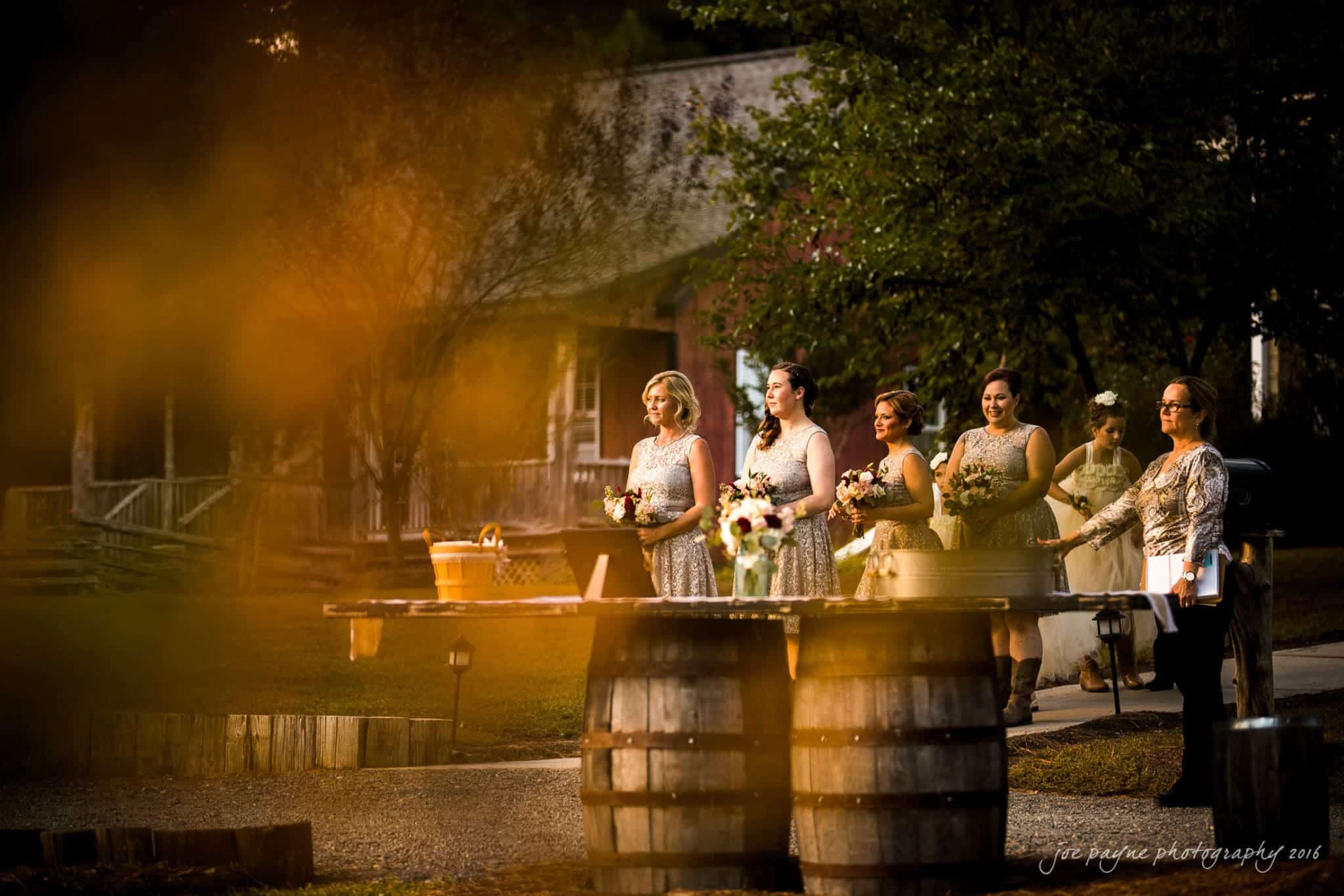 shady wagon farm wedding photography - danielle & josh A bridesmaid procession walks along a rustic path during an outdoor wedding ceremony. They hold bouquets, dressed in matching outfits, with trees and a barn in the background. Sunlight casts a warm glow over the scene.