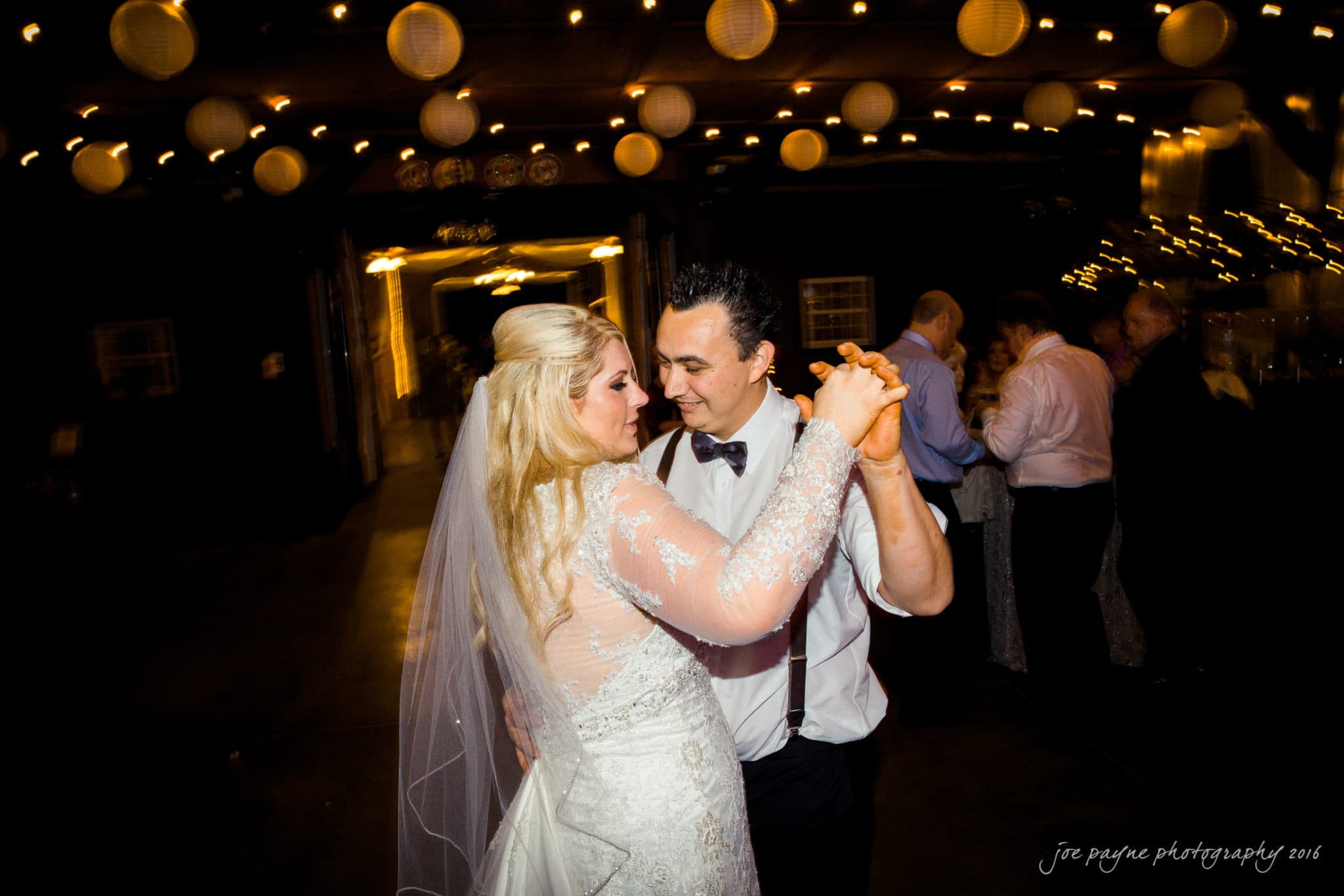 shady wagon farm wedding photography - danielle & josh A bride and groom share a dance under string lights and paper lanterns in a warmly lit venue. The bride wears a lace dress and veil, while the groom is in a shirt with suspenders. Guests are visible in the dimly lit background.