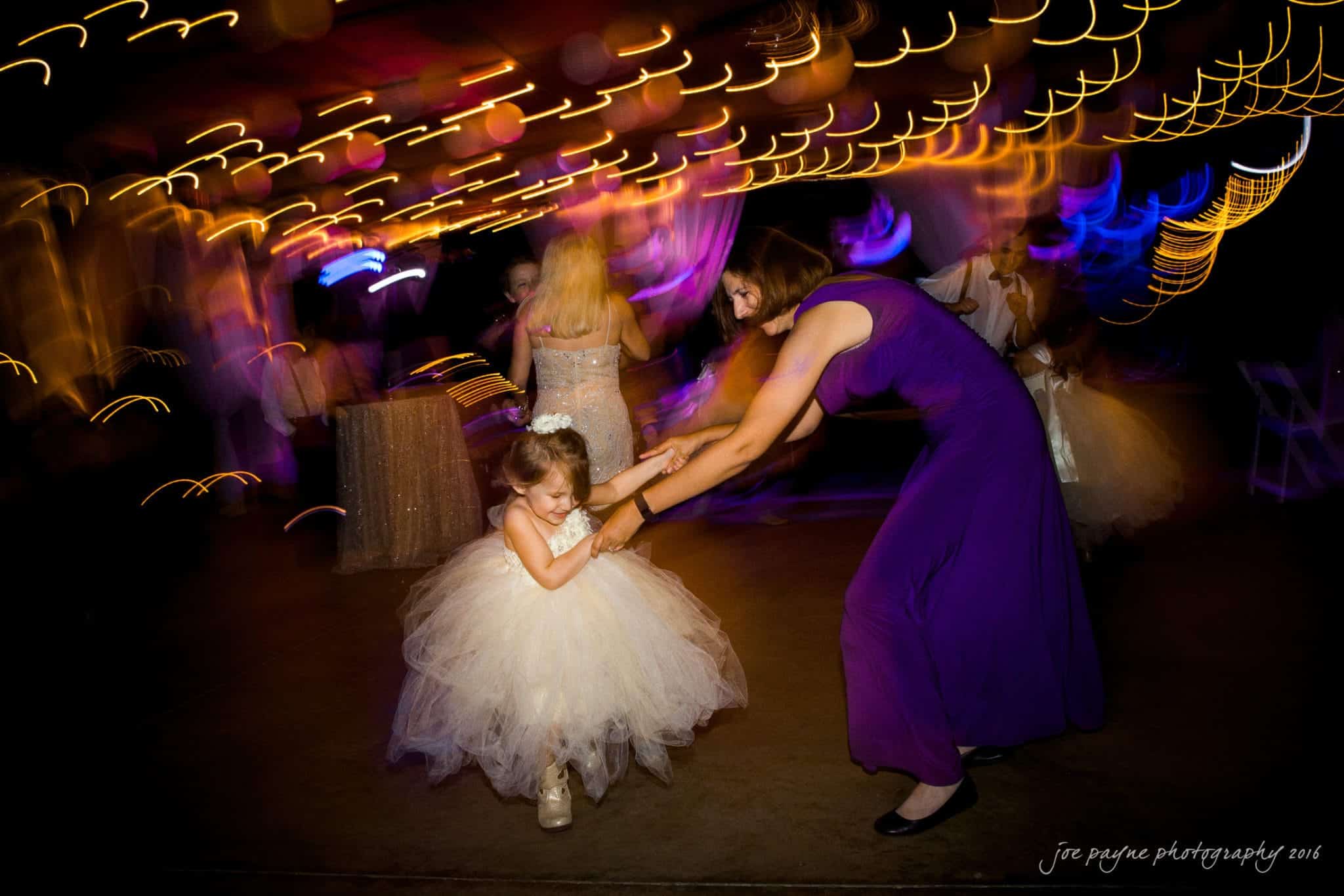 shady wagon farm wedding photography - danielle & josh A woman in a purple dress twirls a young girl in a white, fluffy dress on a dance floor. The background is filled with blurred, colorful lights, creating a lively and festive atmosphere.