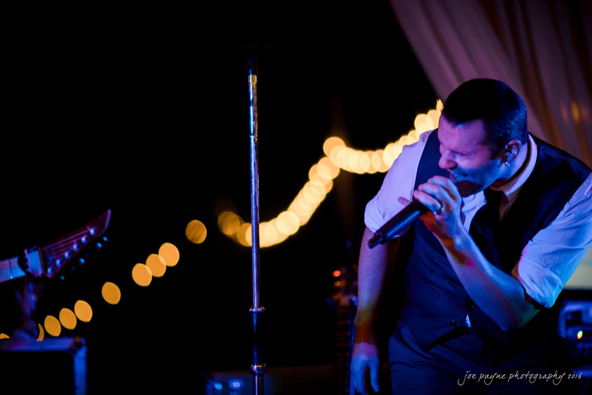 shady wagon farm wedding photography - danielle & josh A singer passionately performs on stage, holding a microphone close to his mouth. Hes wearing a vest over a collared shirt. In the background, warm string lights are strung, creating a cozy ambiance.