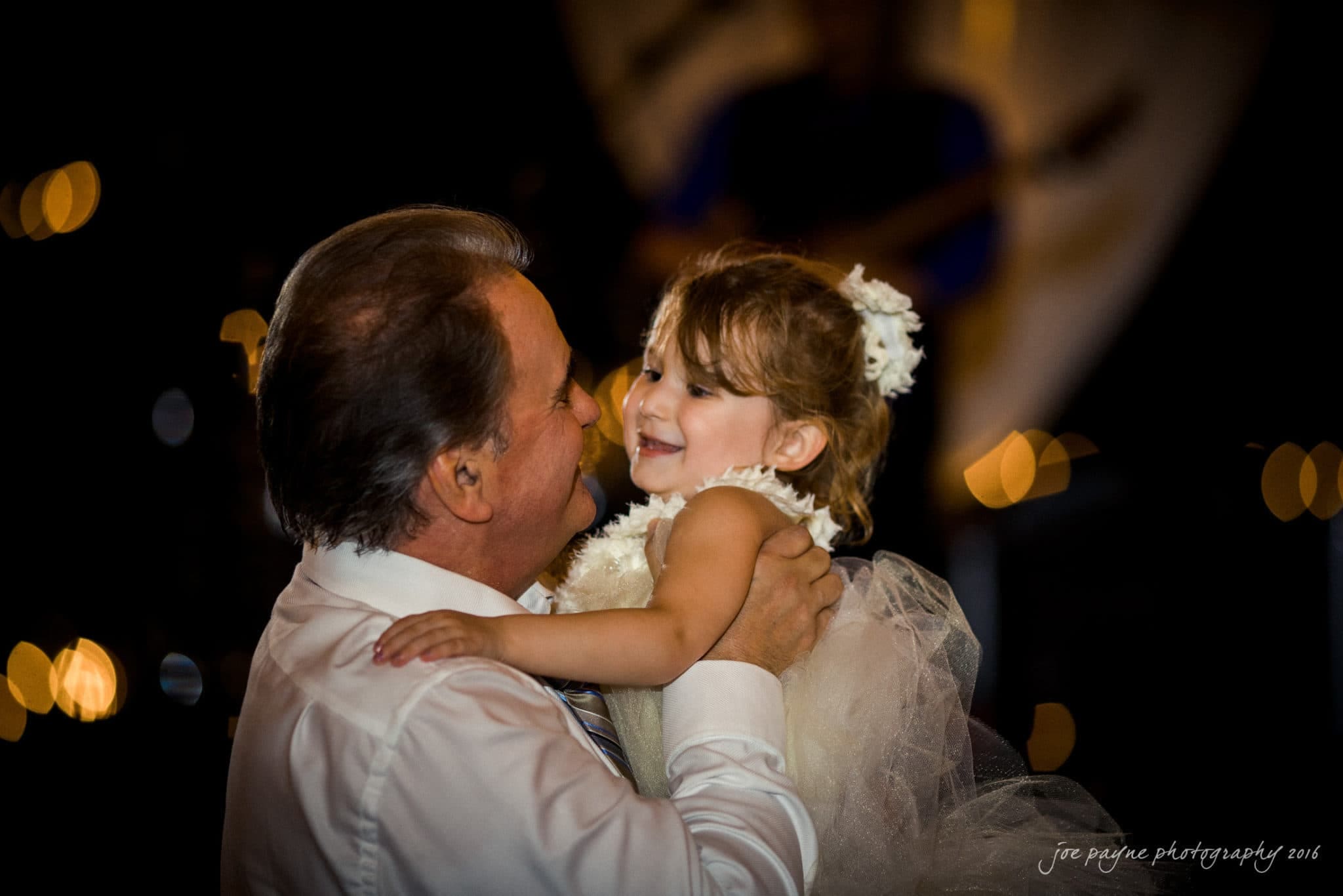 shady wagon farm wedding photography - danielle & josh A man in a white shirt holds a smiling young girl in a white dress. They are dancing together in a warmly lit, festive setting with blurred lights in the background.
