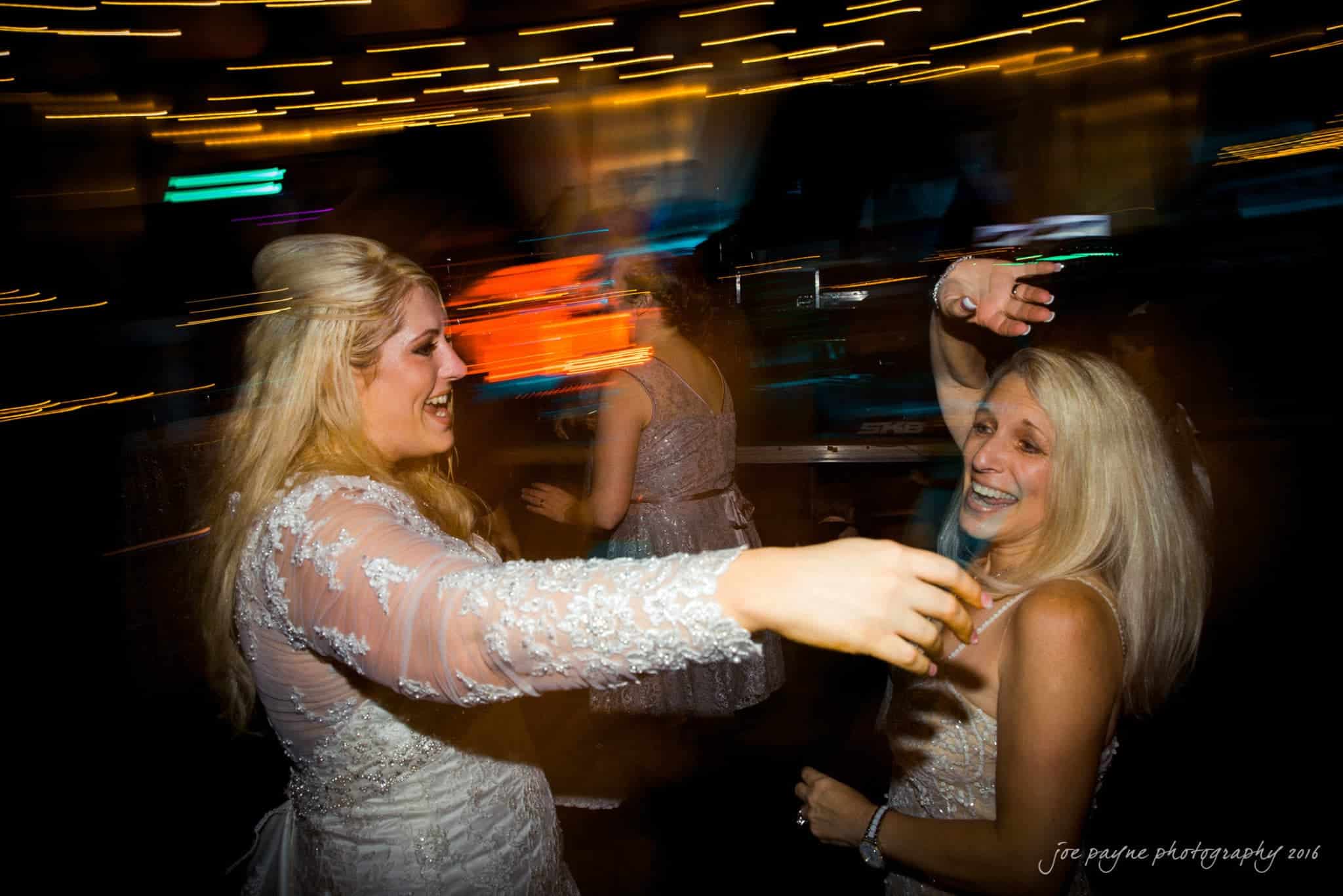 shady wagon farm wedding photography - danielle & josh Two women dressed in elegant attire dance joyfully at a lively event, surrounded by blurred lights. One woman extends her arm while the other laughs with a raised arm. The atmosphere is vibrant and festive.