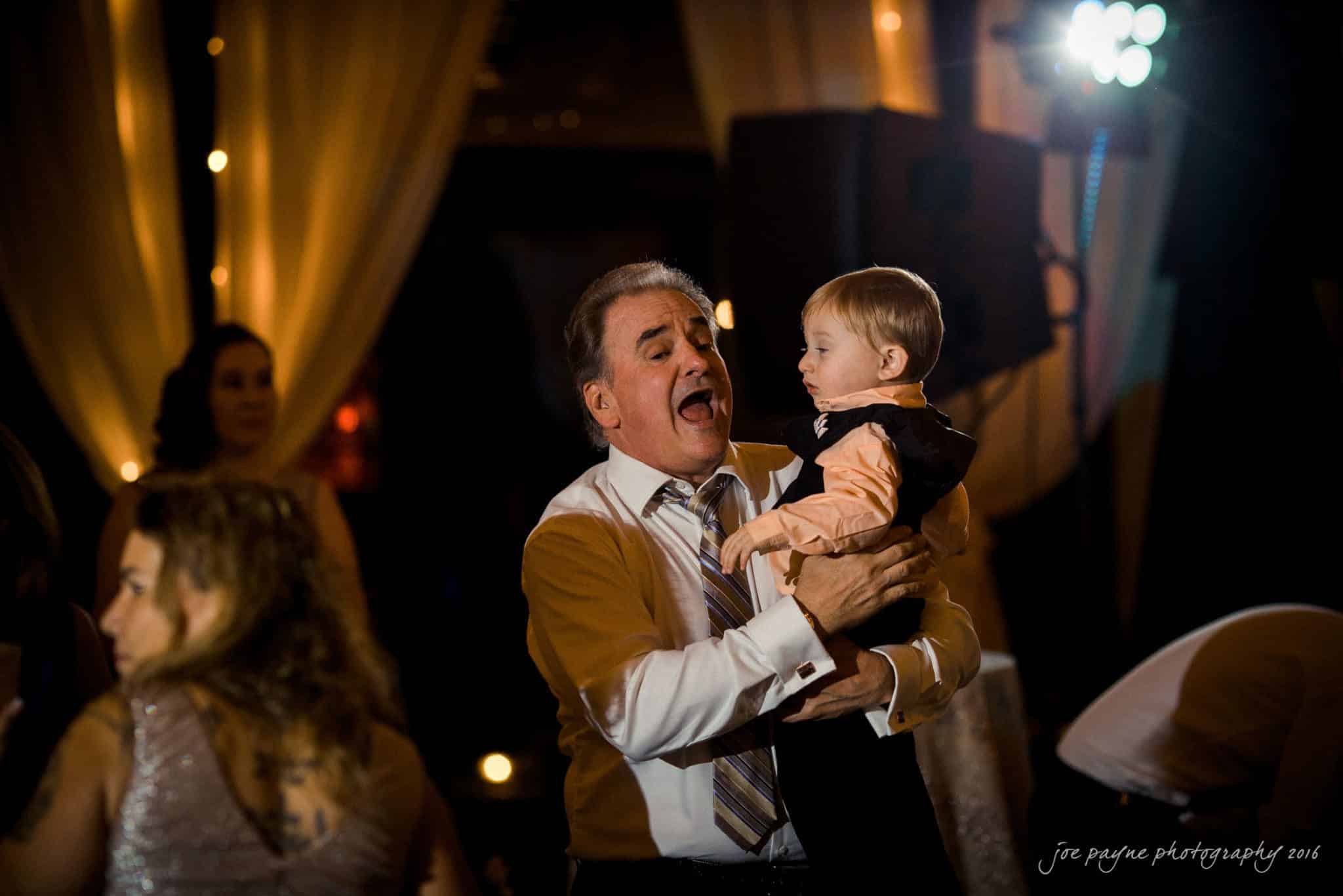 shady wagon farm wedding photography - danielle & josh A man in a dress shirt and tie joyfully dances while holding a young child in a suit. They are surrounded by softly lit decorations, including draped curtains and dim lights, creating a festive atmosphere.