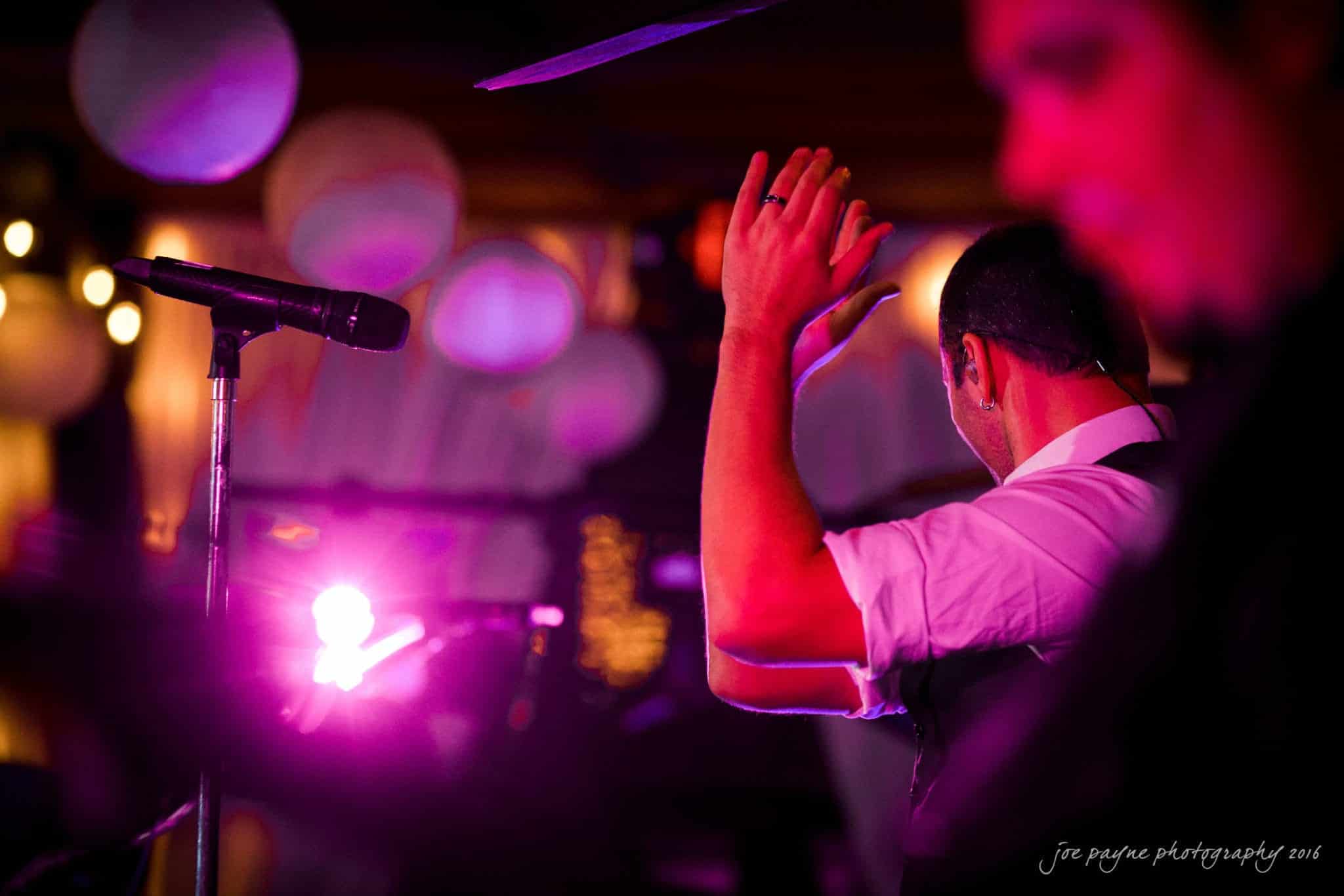 shady wagon farm wedding photography - danielle & josh A performer claps on stage under pink and purple lights, with a microphone nearby. The background is softly blurred with colorful bokeh effects, and another persons silhouette is visible in the foreground.