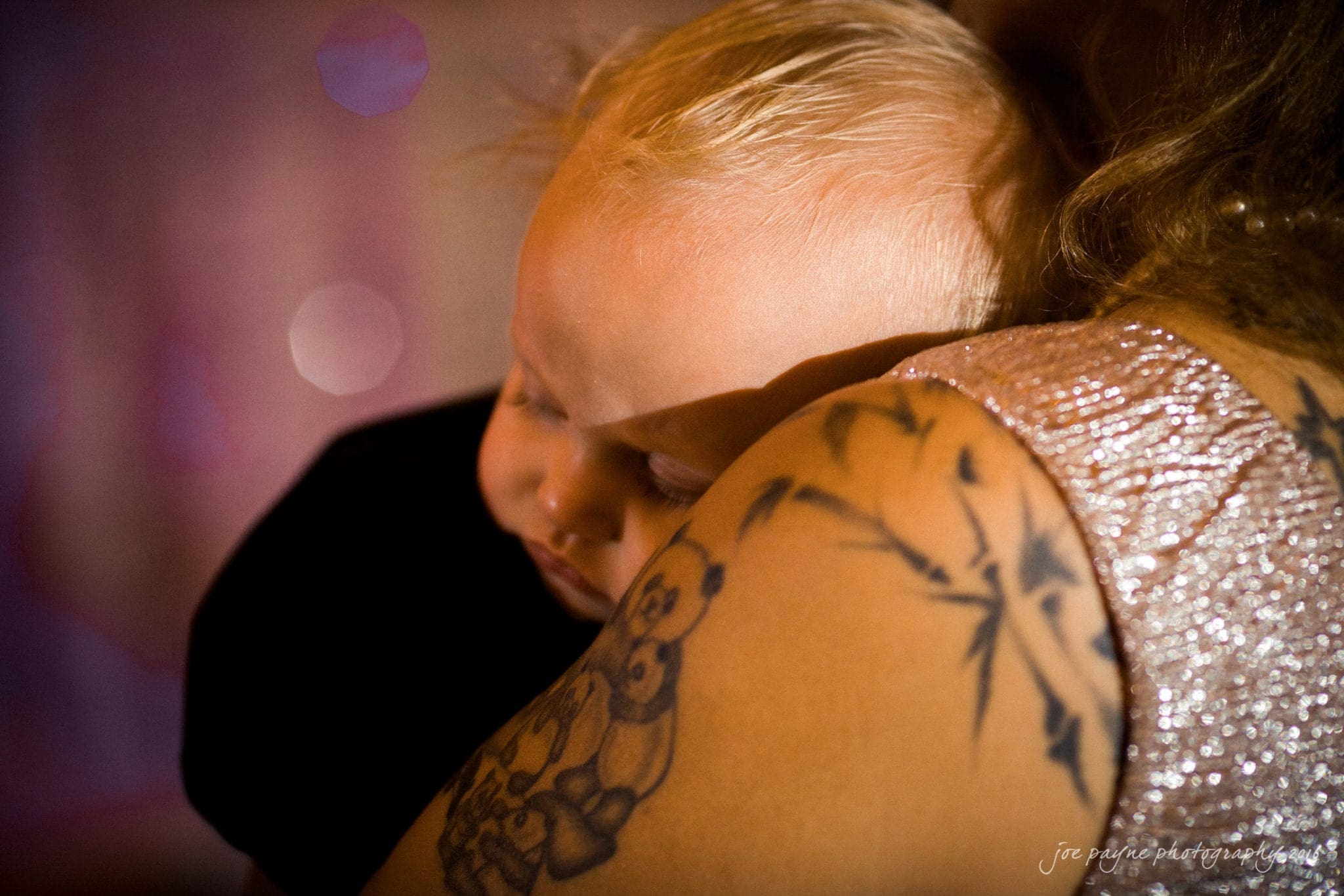 shady wagon farm wedding photography - danielle & josh A baby with blonde hair sleeps peacefully on a persons shoulder. The person is wearing a sparkly top and has a prominent tattoo featuring a panda and bamboo leaves on their arm. The background is softly lit with pinkish tones.