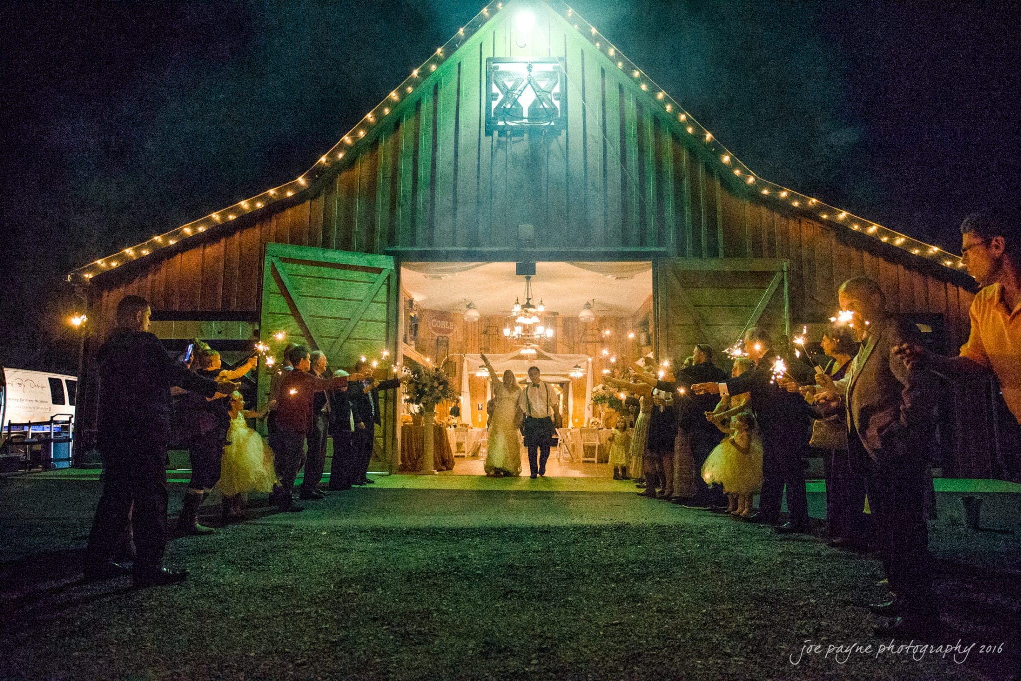 shady wagon farm wedding photography - danielle & josh A couple walks hand in hand out of a barn at night, surrounded by people holding sparklers. The barn is warmly lit, with wooden doors wide open and string lights along the roof.