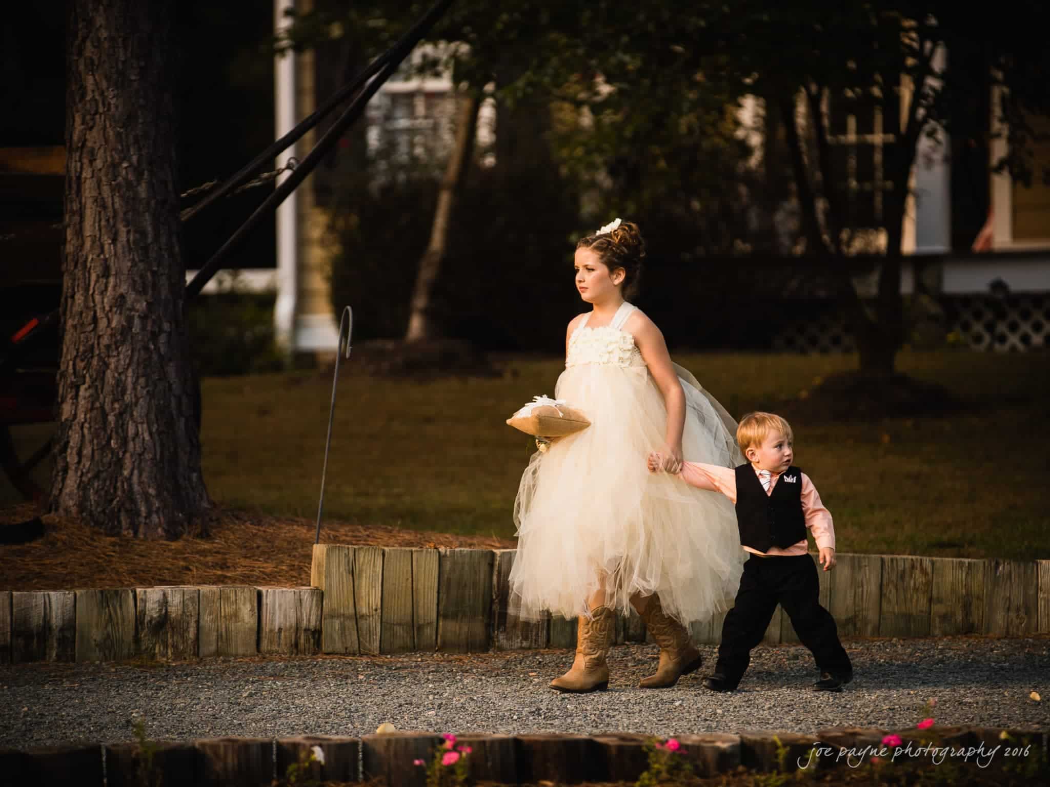 shady wagon farm wedding photography - danielle & josh A young girl in a white, fluffy dress and boots holds hands with a young boy in formal attire. They walk on a gravel path with trees and a house in the background, captured in warm lighting.