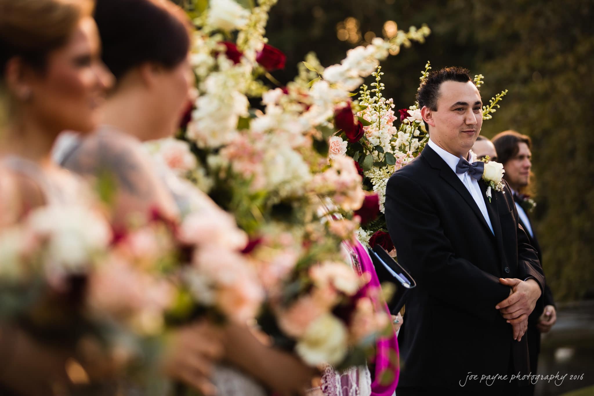 shady wagon farm wedding photography - danielle & josh A groom stands smiling at an outdoor wedding ceremony, dressed in a black suit and bowtie, surrounded by lush floral arrangements. Bridesmaids in soft dresses hold bouquets, and greenery is visible in the background.