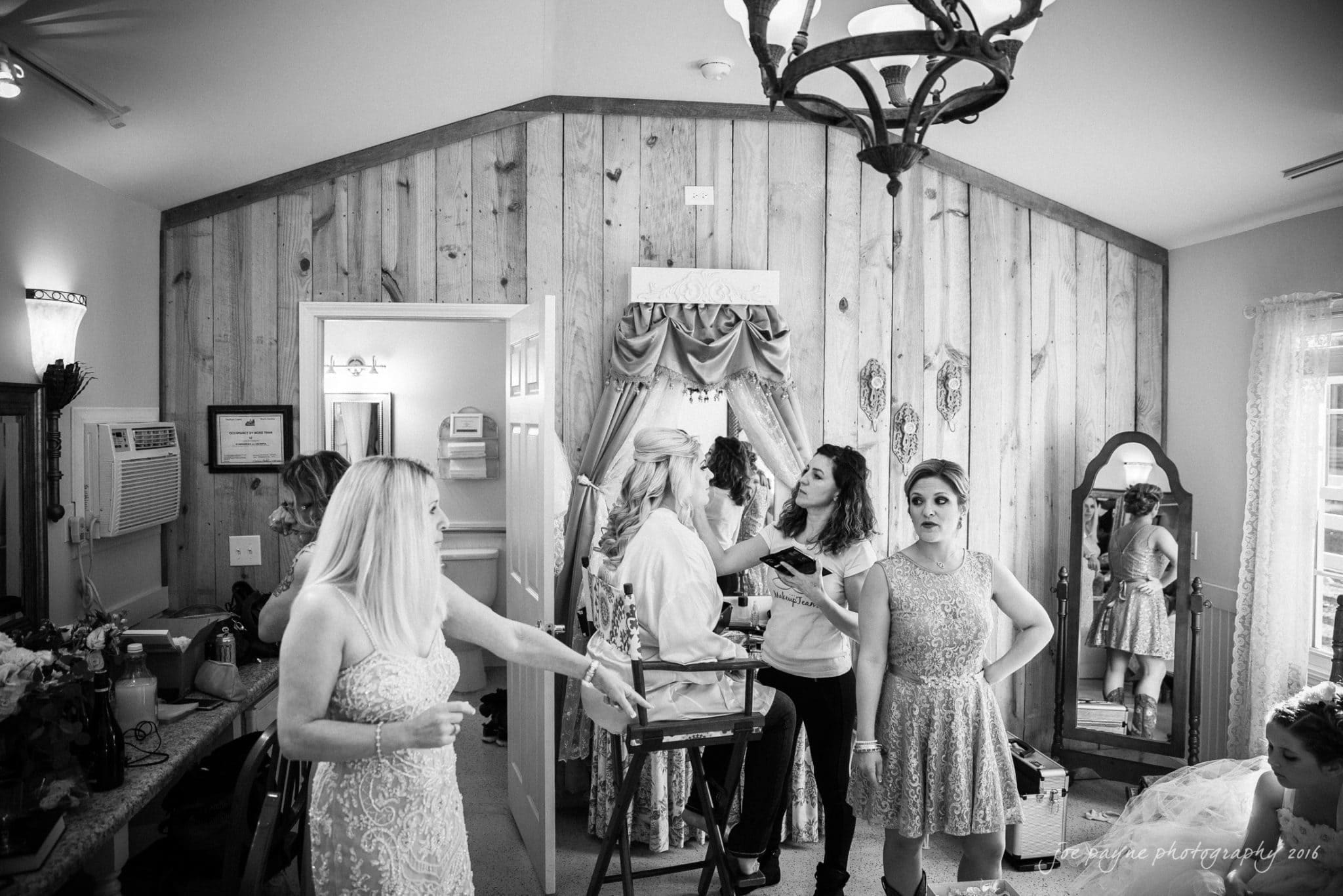 shady wagon farm wedding photography - danielle & josh Black and white photo of a bridal party getting ready in a room with wood paneled walls. Women are chatting and preparing, with one woman standing in the foreground holding a drink and another seated in front of a mirror.