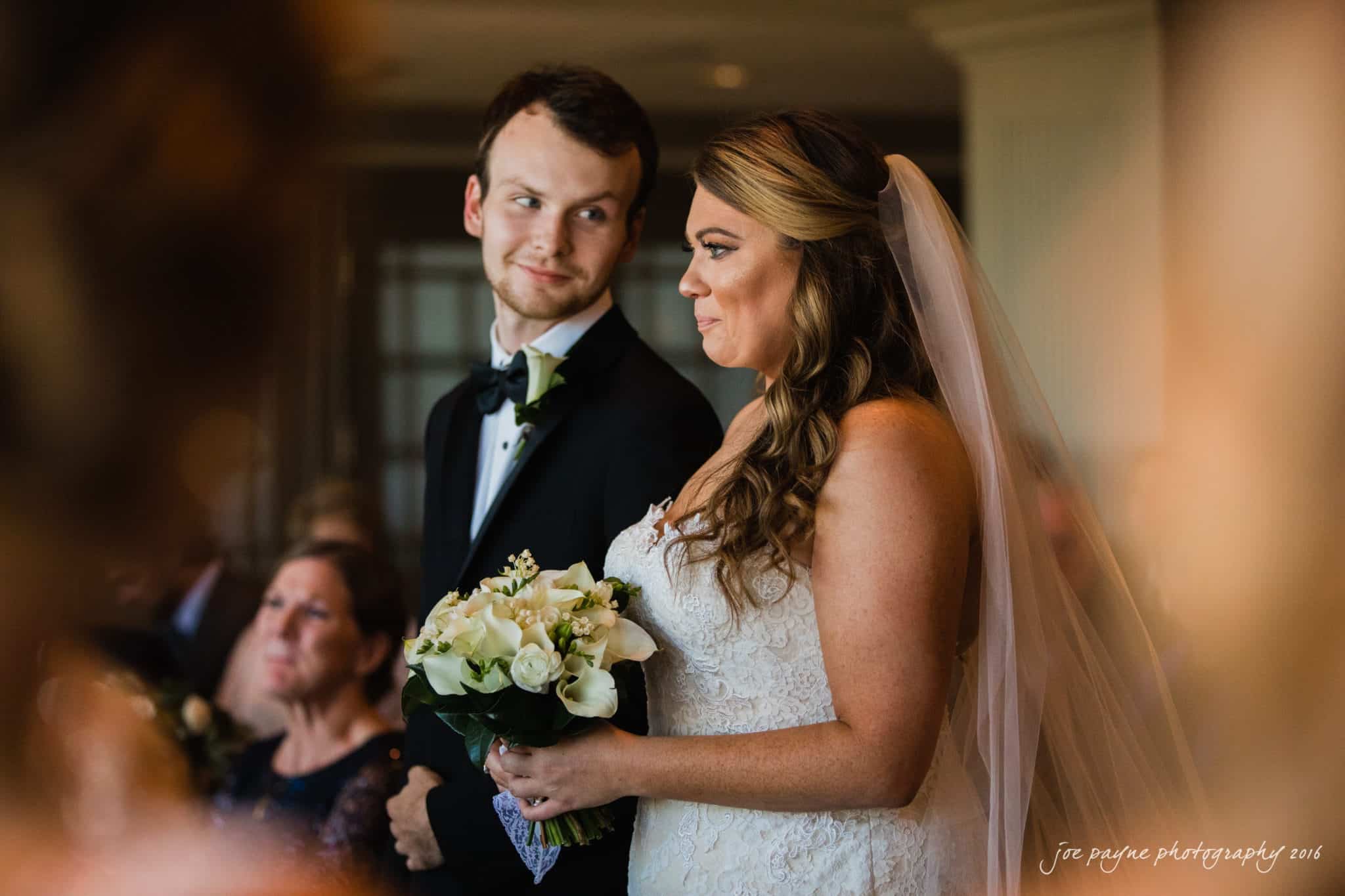 city club wedding photographer - lauren & adam A bride in a white gown and veil holds a bouquet of white flowers while standing beside a groom in a black tuxedo. They are indoors, appearing to be at a wedding ceremony, with guests seated in the background.