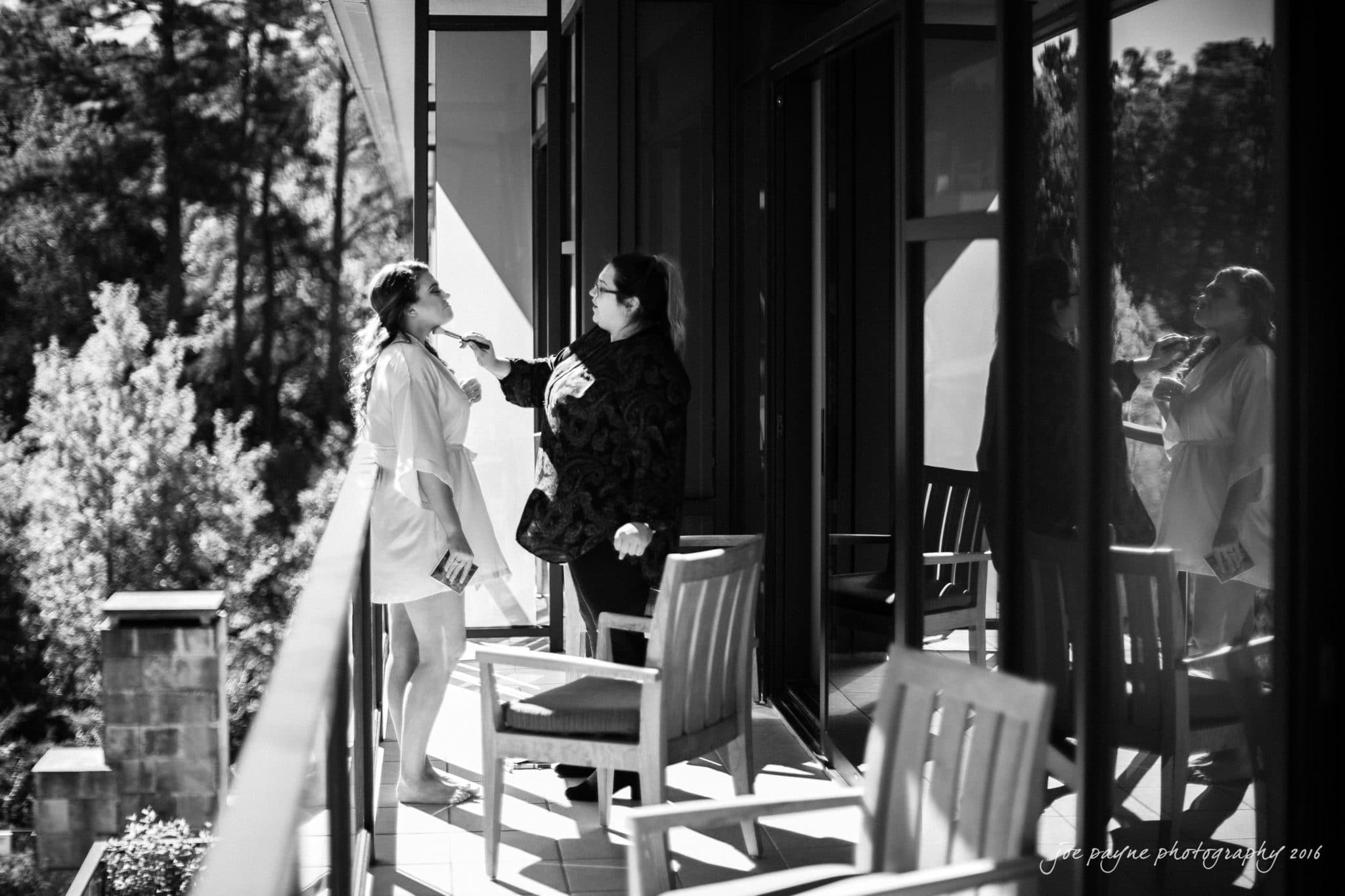 city club wedding photographer - lauren & adam A woman in a robe is standing on a balcony, having makeup applied by another person. The scene is in black and white, with wooden chairs nearby and a view of trees in the background.
