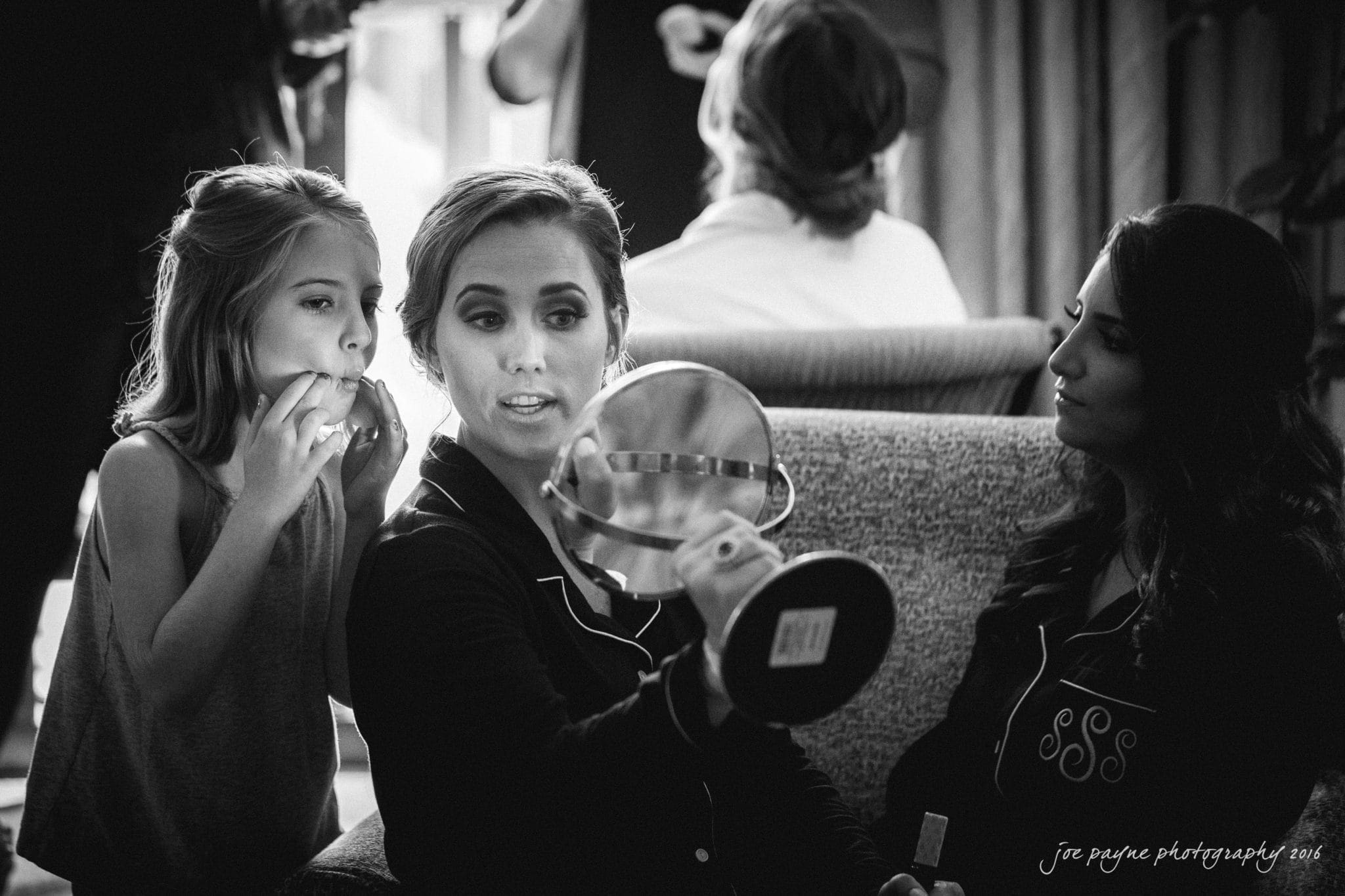 city club wedding photographer - lauren & adam A black and white photo of a woman holding a mirror while applying makeup, with a young girl beside her mimicking the action. Another woman sits nearby, observing. The setting appears relaxed and casual.