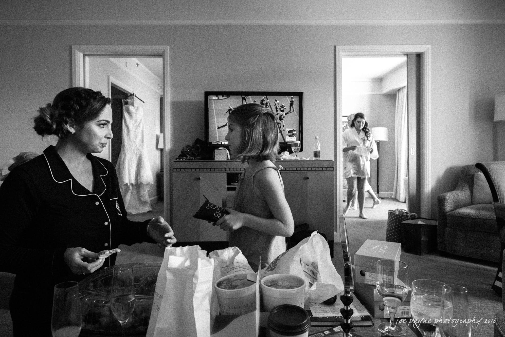city club wedding photographer - lauren & adam A black and white photo of a room with a woman in pajamas talking to a child holding a snack. In the background, another woman in a robe walks through a doorway. The room is cluttered with various items and a TV is on.