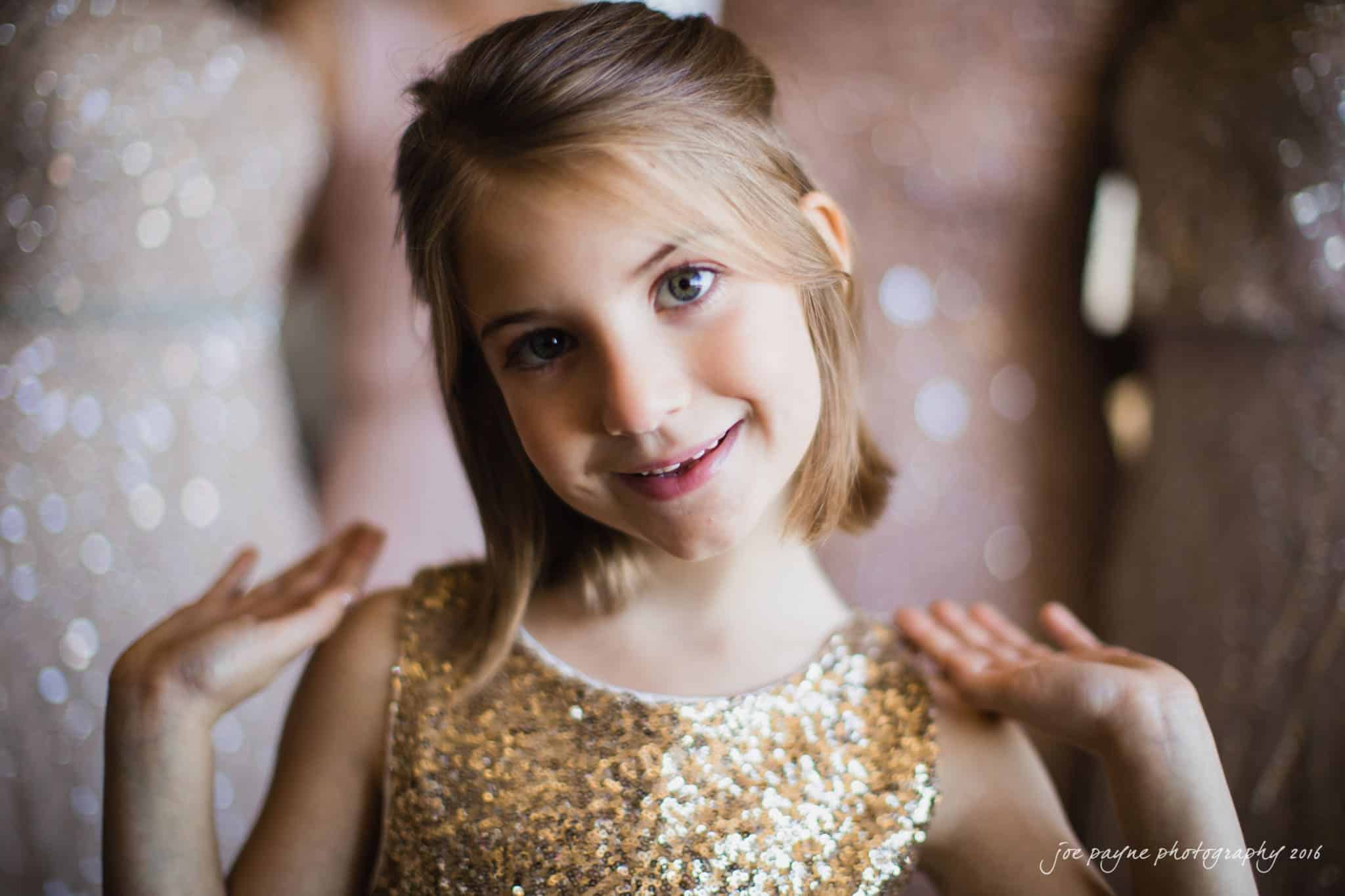 city club wedding photographer - lauren & adam A young girl with shoulder-length hair smiles at the camera, wearing a sparkling gold dress. Her surroundings are softly blurred, highlighting the glittery texture of her outfit.