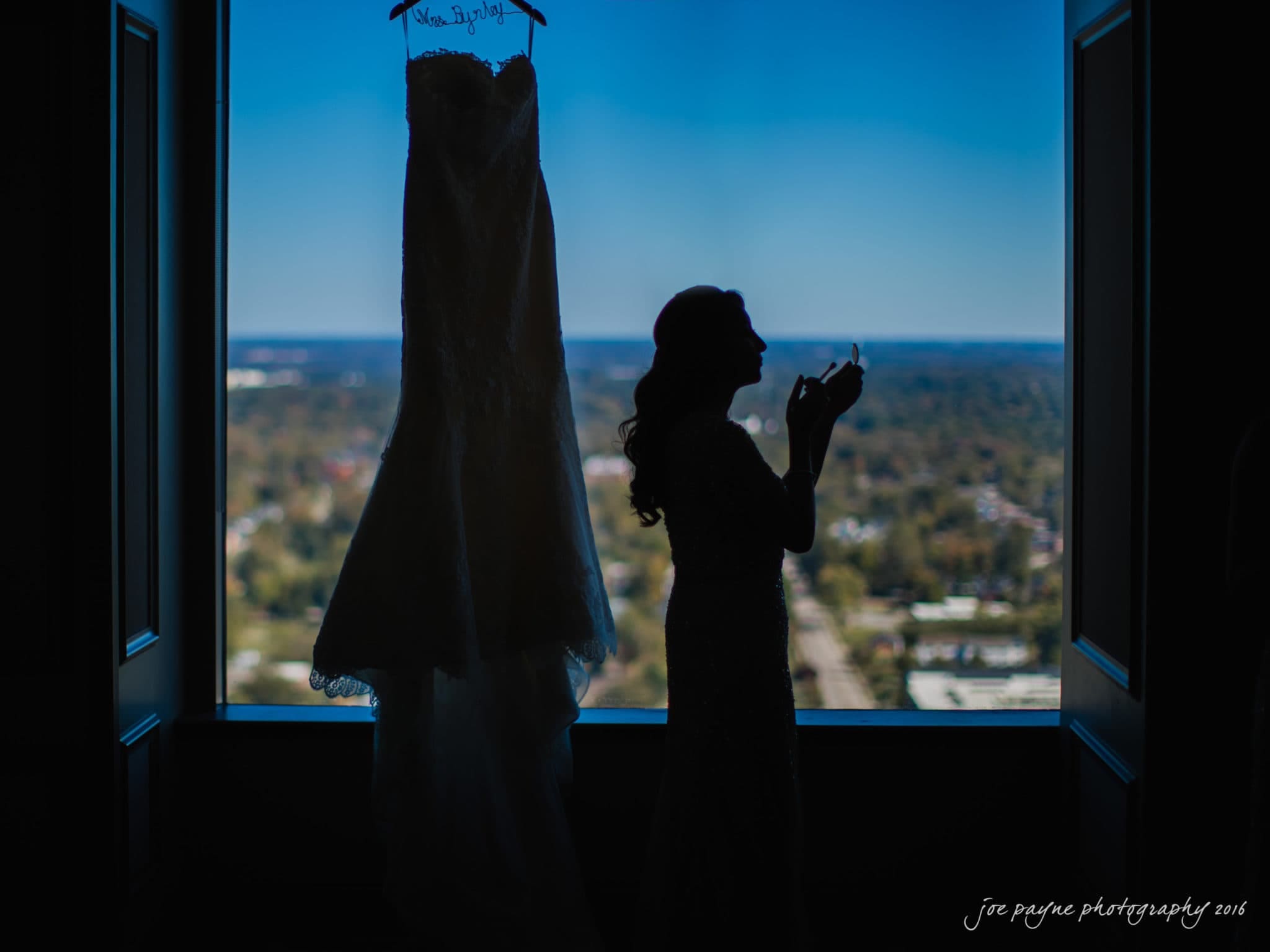city club wedding photographer - lauren & adam Silhouette of a woman applying makeup, standing beside a window that overlooks a scenic landscape and features a hanging wedding dress.