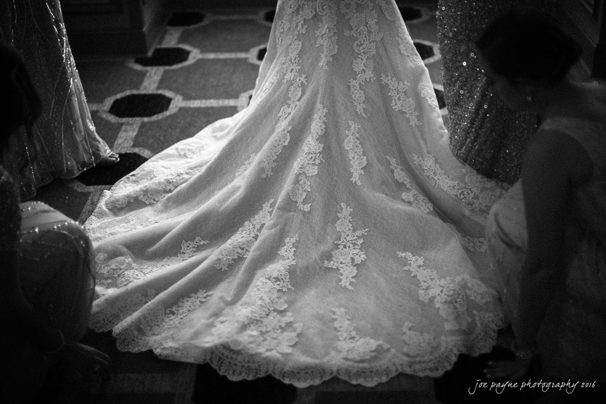 city club wedding photographer - lauren & adam A black and white image of a brides lace wedding gown with a long train elegantly spread on the floor. Three women are gathered around, adjusting the dress. The intricate lace pattern is prominently displayed.