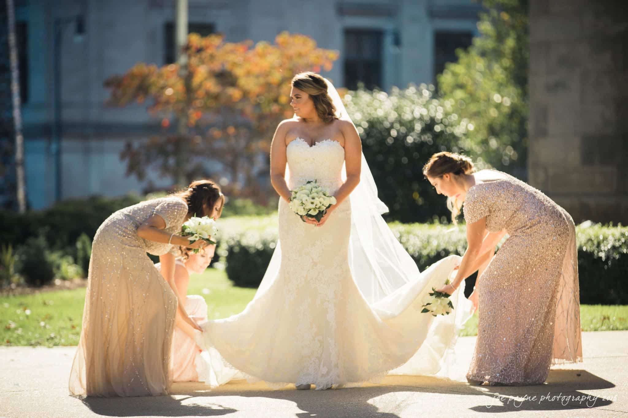city club wedding photographer - lauren & adam A bride in a strapless wedding gown stands outdoors holding flowers. Two bridesmaids adjust her dress train on either side. Sunlight filters through trees in the background, creating a warm, picturesque scene.
