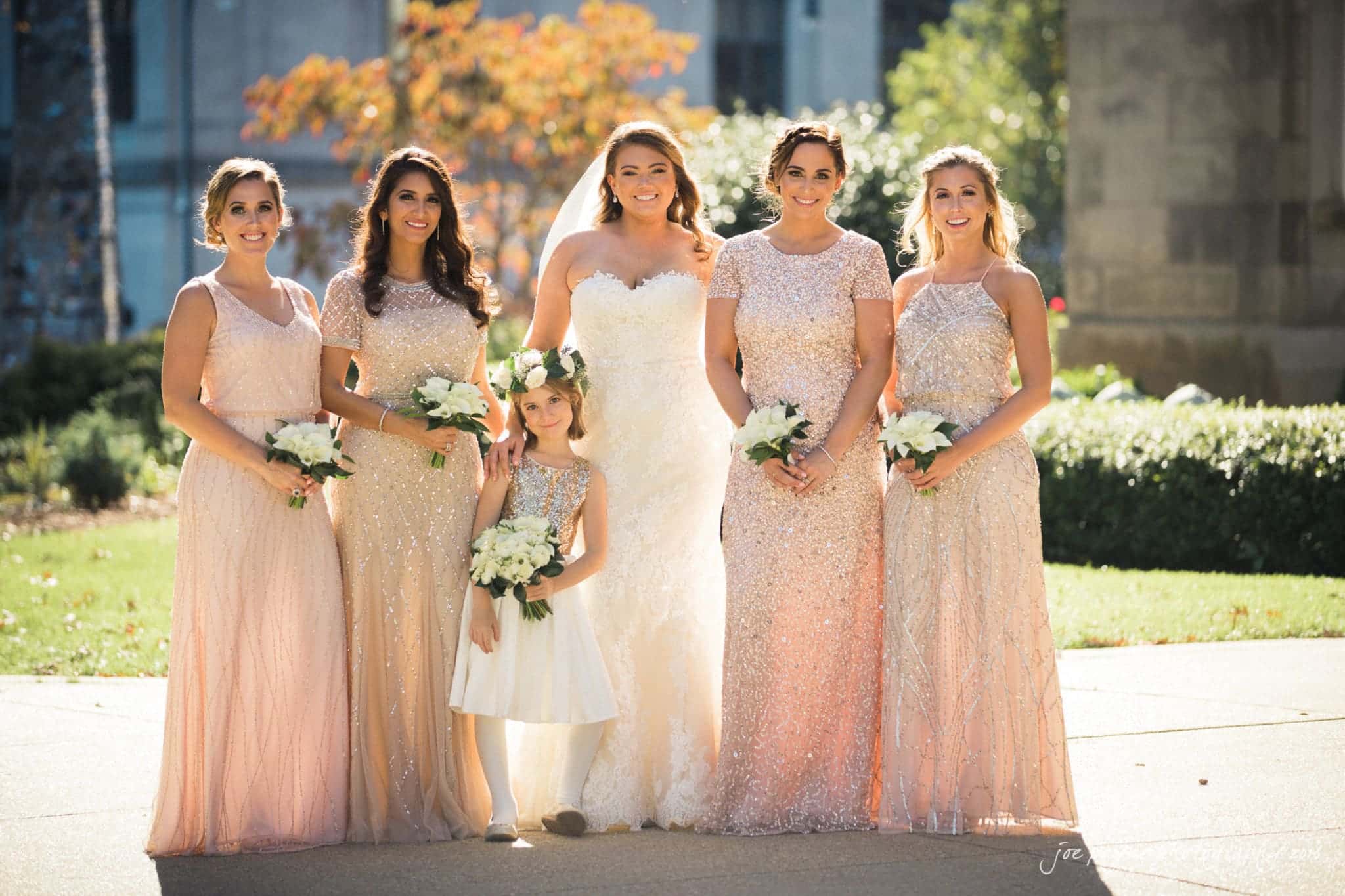 city club wedding photographer - lauren & adam A bride and five bridesmaids stand outdoors in elegant gowns, holding white flower bouquets. The young girl in the front wears a white dress and flower crown. Sunlight filters through trees, casting a warm glow on the group.