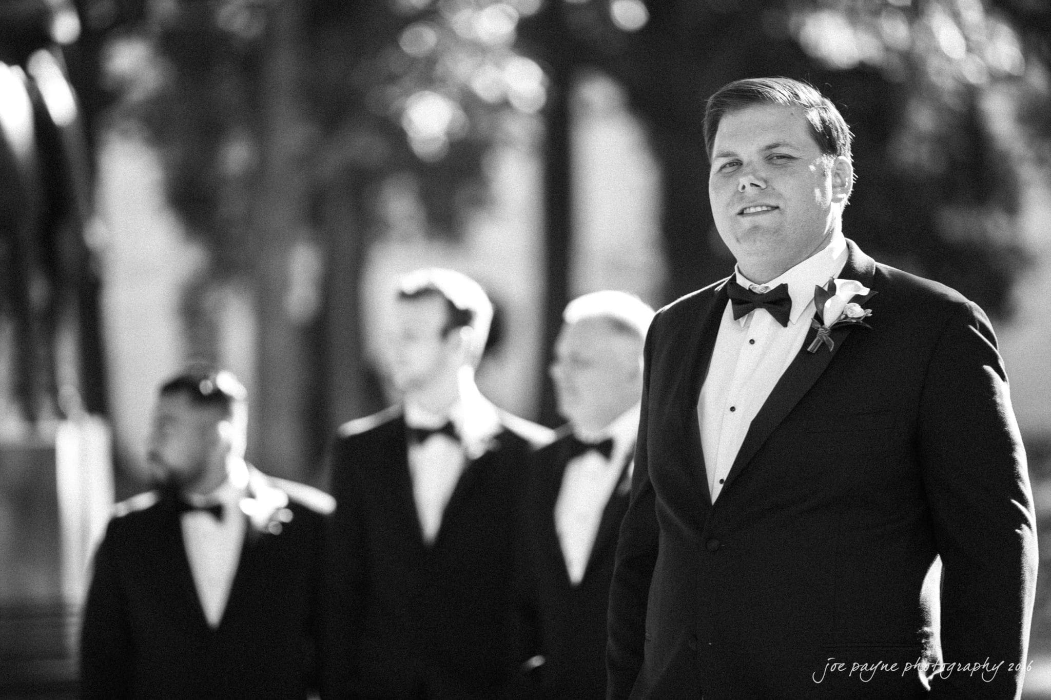 city club wedding photographer - lauren & adam A black and white photo of a groom in a tuxedo with a boutonniere, standing outside. Three groomsmen in tuxedos stand behind him, slightly out of focus. Trees and a statue are visible in the background.
