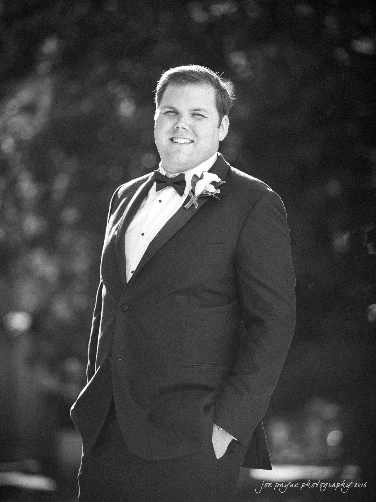 city club wedding photographer - lauren & adam A man in a tuxedo stands outdoors, smiling with one hand in his pocket. The background is blurred, highlighting him as the focal point of the black-and-white photo.