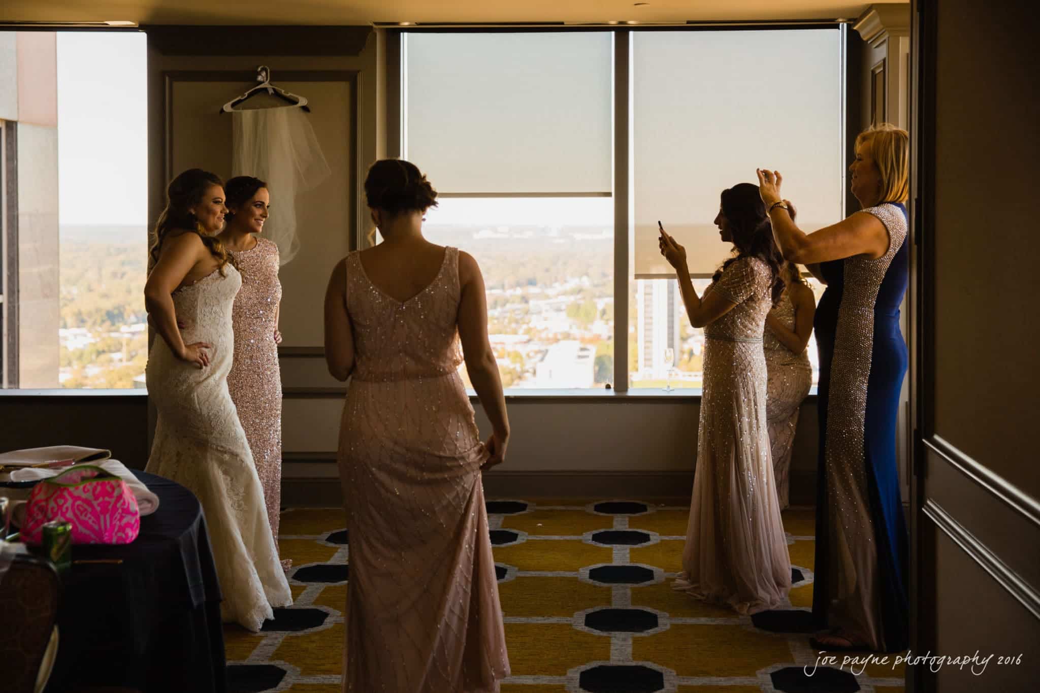 city club wedding photographer - lauren & adam A bride and bridesmaids in elegant dresses stand in a hotel room with city views. One woman holds a phone, capturing a photo. The scene is filled with soft, natural light from the large window.