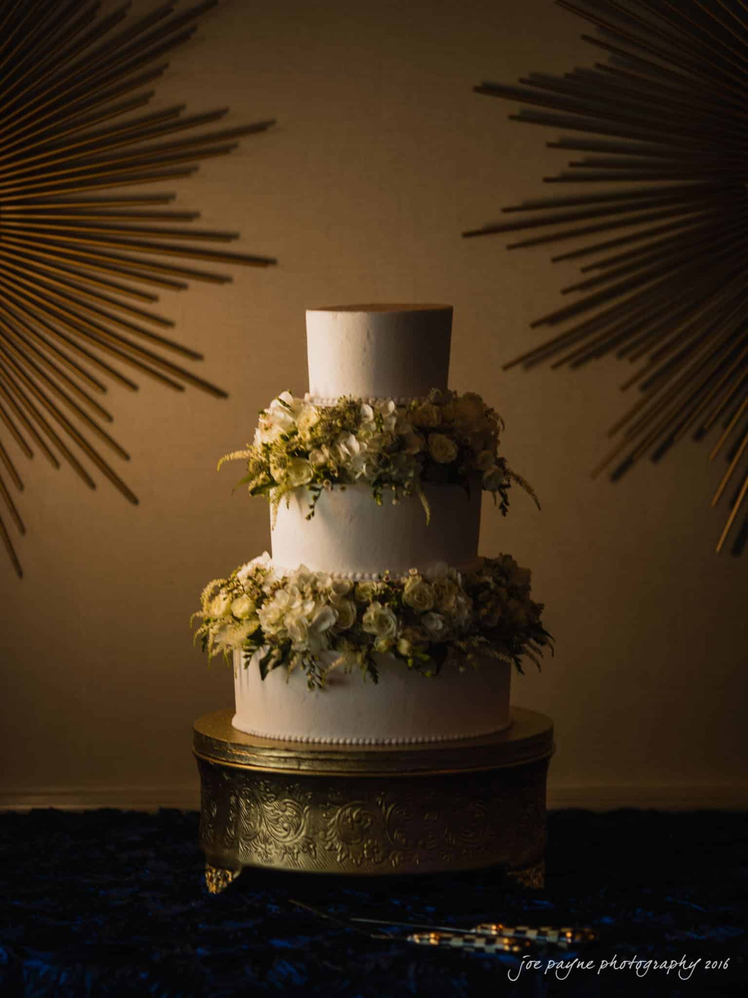 city club wedding photographer - lauren & adam Three-tier white wedding cake adorned with white roses and greenery, placed on an ornate golden stand. The backdrop features sunburst decorations on a textured wall.