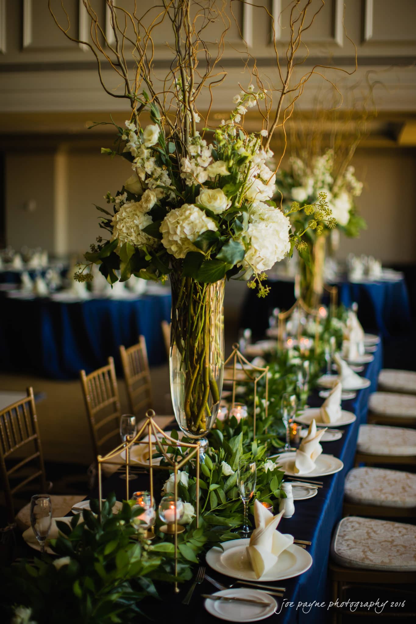 city club wedding photographer - lauren & adam Elegant dining table setting with tall vases of white flowers and greenery. The table features candles in geometric holders and neatly arranged tableware. Soft lighting creates a warm, inviting ambiance, with navy tablecloths and chiavari chairs.
