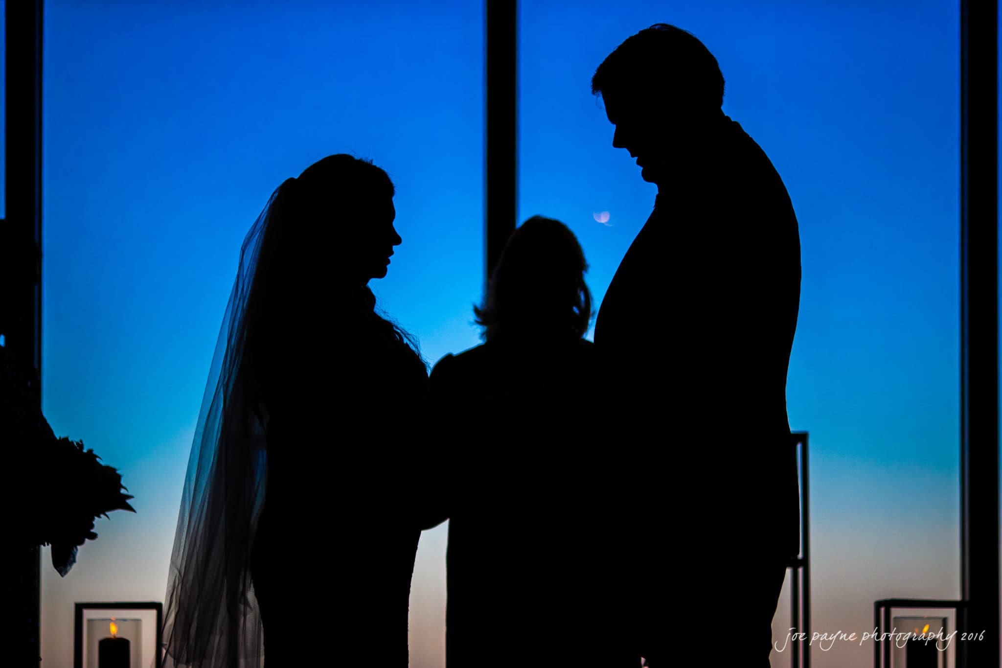 city club wedding photographer - lauren & adam Silhouetted against a deep blue sky, a bride and groom face each other, accompanied by a third person between them. The scene is illuminated by a shimmering crescent moon, creating a romantic and serene atmosphere.