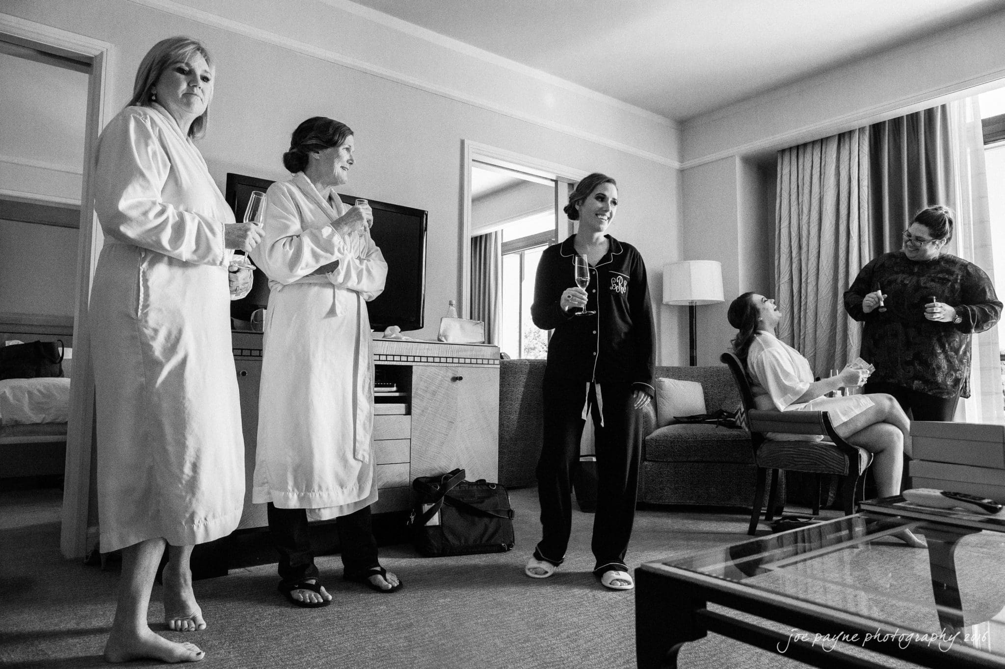 city club wedding photographer - lauren & adam A group of people in casual attire gather in a hotel room. Two stand in robes, two are seated near a window with curtains drawn, and one is in loungewear. They seem relaxed, engaging in conversation, with a television in the background.