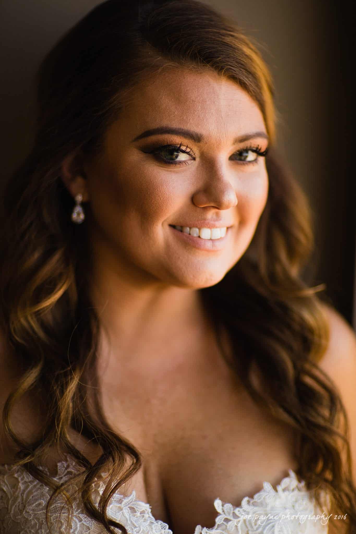 city club wedding photographer - lauren & adam A woman with long, wavy brown hair is smiling. She is wearing a white lace top and pearl drop earrings. The lighting is warm and soft, highlighting her makeup and features.