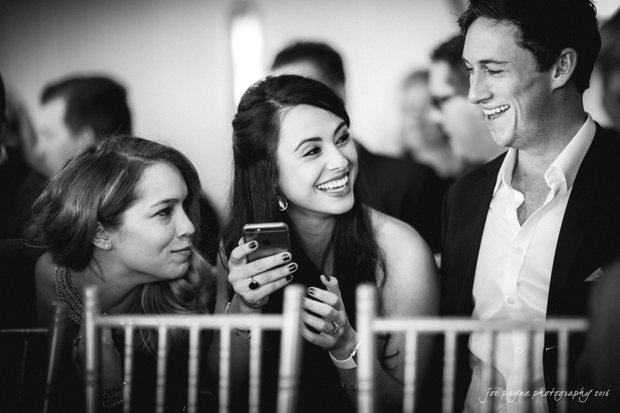 city club wedding photographer - lauren & adam Three people in formal attire are smiling and laughing together. One woman is holding a phone, and they are seated behind wooden chairs. The image is black and white, capturing a candid, joyful moment.
