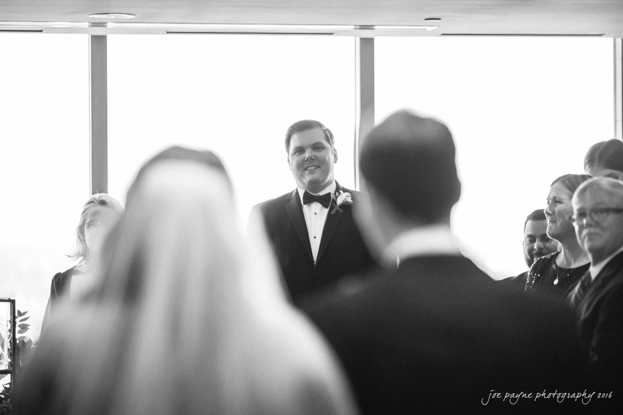 city club wedding photographer - lauren & adam A bride with long hair walks down the aisle towards a groom in a tuxedo, accompanied by guests sitting and standing on either side, in a bright wedding venue with large windows. The groom is smiling as he watches her approach.
