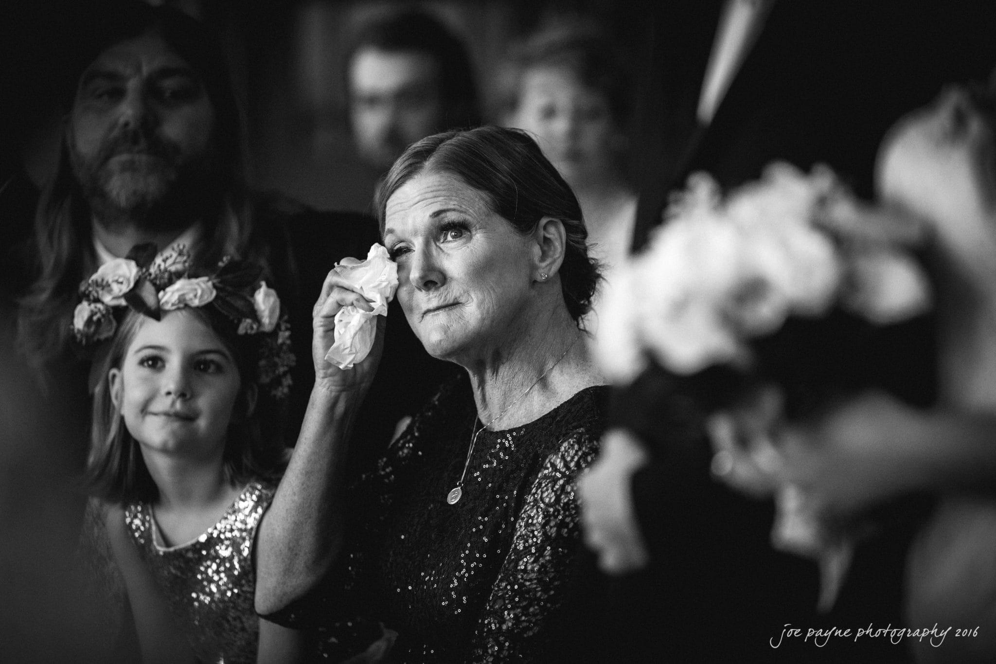 city club wedding photographer - lauren & adam A woman in a dark dress wipes tears from her eyes with a tissue at a formal event. A young girl in a sequined dress and floral headband looks on. Other people are blurred in the background.