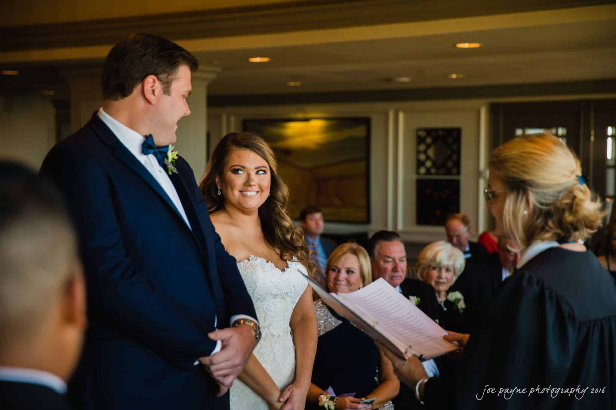 city club wedding photographer - lauren & adam A bride and groom smile at each other during a wedding ceremony. The officiant stands to the right, holding a book. Guests are seated in the background, attentively watching the couple. The atmosphere is warm and joyful.