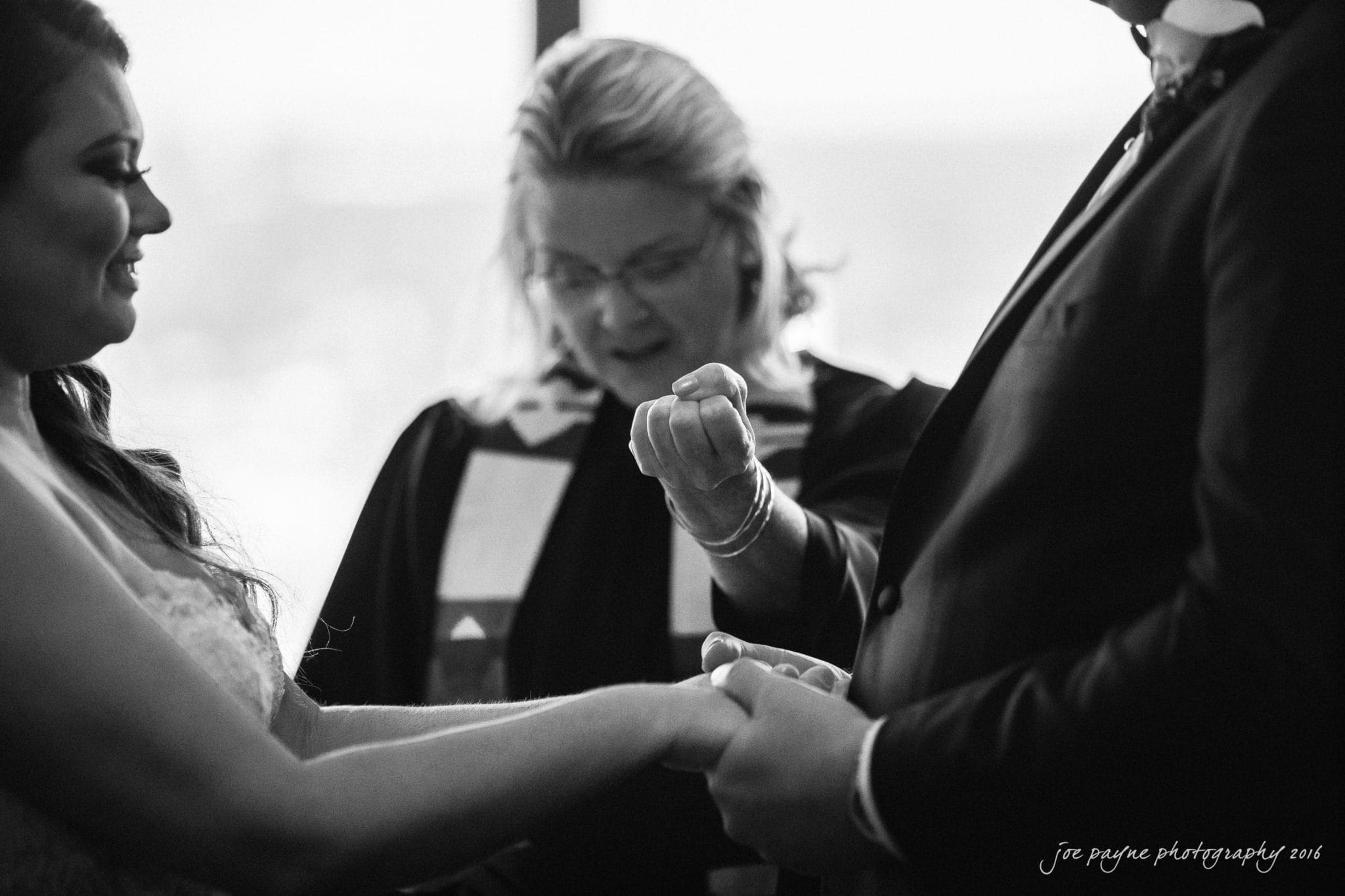 city club wedding photographer - lauren & adam A black-and-white photo of a wedding ceremony. The bride and groom stand facing each other, holding hands. An officiant behind them gestures with one hand while holding a script. The atmosphere is solemn and intimate.