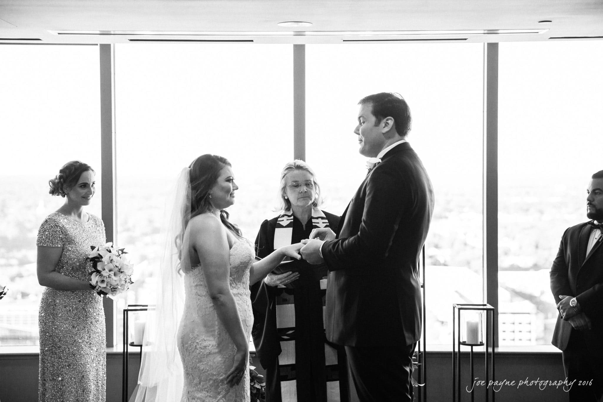 city club wedding photographer - lauren & adam A bride and groom stand facing each other during their wedding ceremony. The officiant, wearing a stole, stands between them. A bridesmaid holds flowers to the left. The background shows a bright cityscape through large windows.