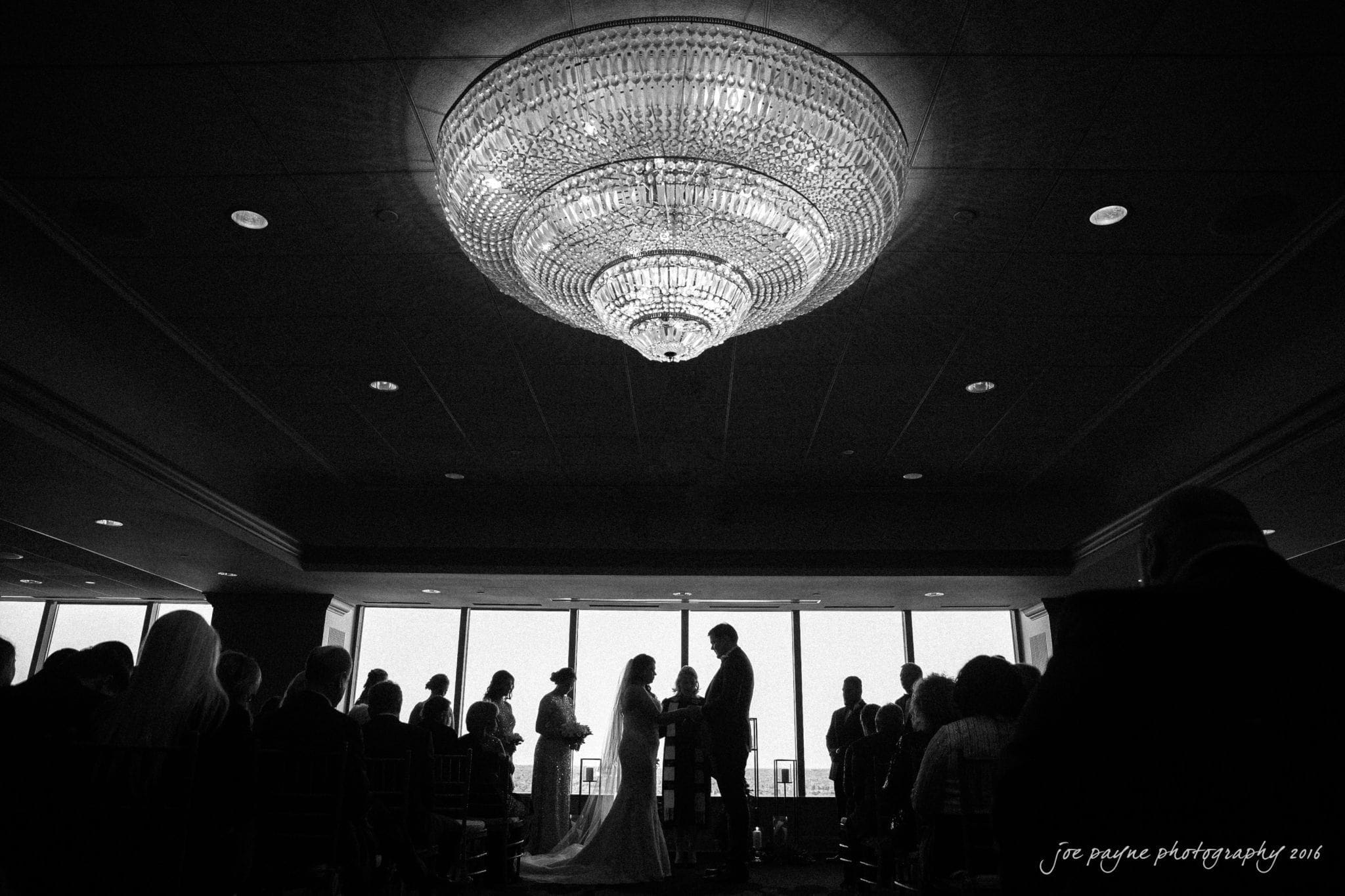 city club wedding photographer - lauren & adam A black and white photo of a wedding ceremony. The bride and groom stand facing each other, silhouetted against a large window. Guests are seated on both sides. A grand chandelier hangs above, illuminating the scene.