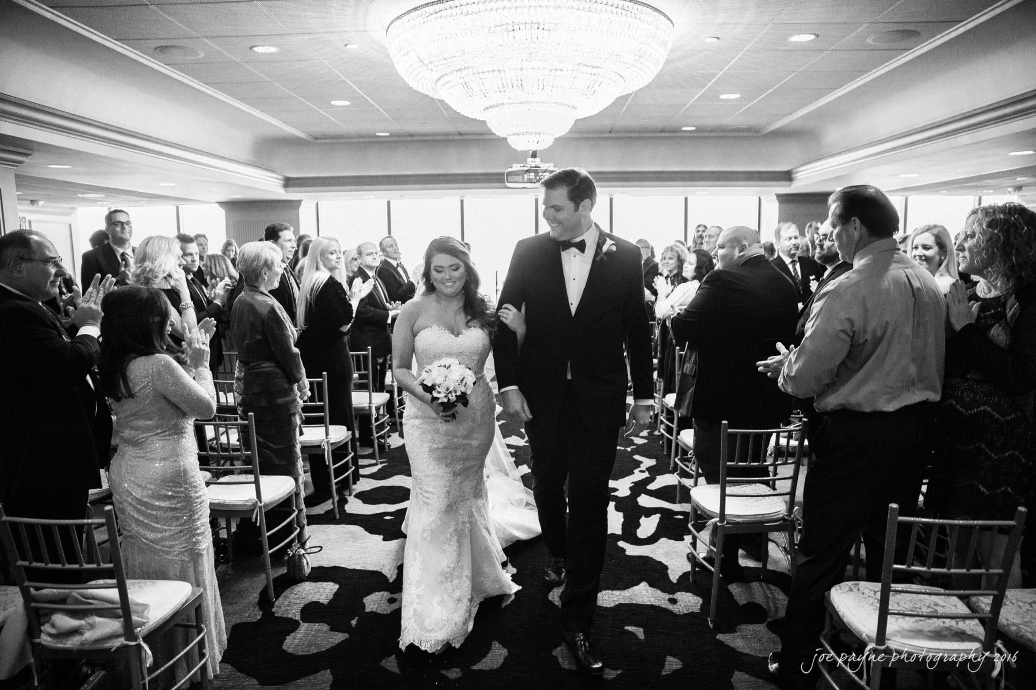 city club wedding photographer - lauren & adam A bride and groom walk down the aisle together in a wedding ceremony. Guests are standing and clapping in a decorated venue with a large chandelier overhead. The bride holds a bouquet, and both are smiling. The image is in black and white.