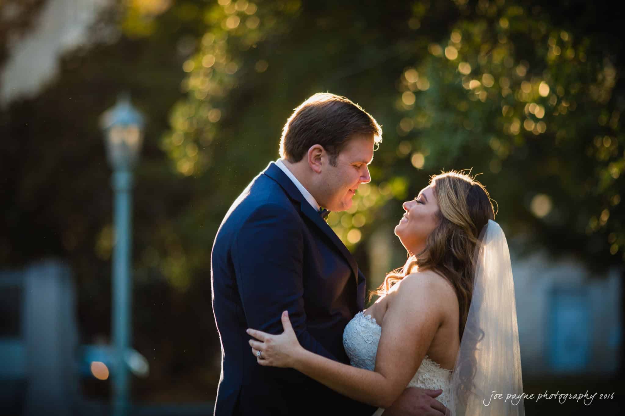 city club wedding photographer - lauren & adam A bride and groom embrace outdoors during their wedding. Sunlight filters through trees, casting a warm glow around them. Both are smiling and gazing into each others eyes, with the bride wearing a veil and the groom in a dark suit.