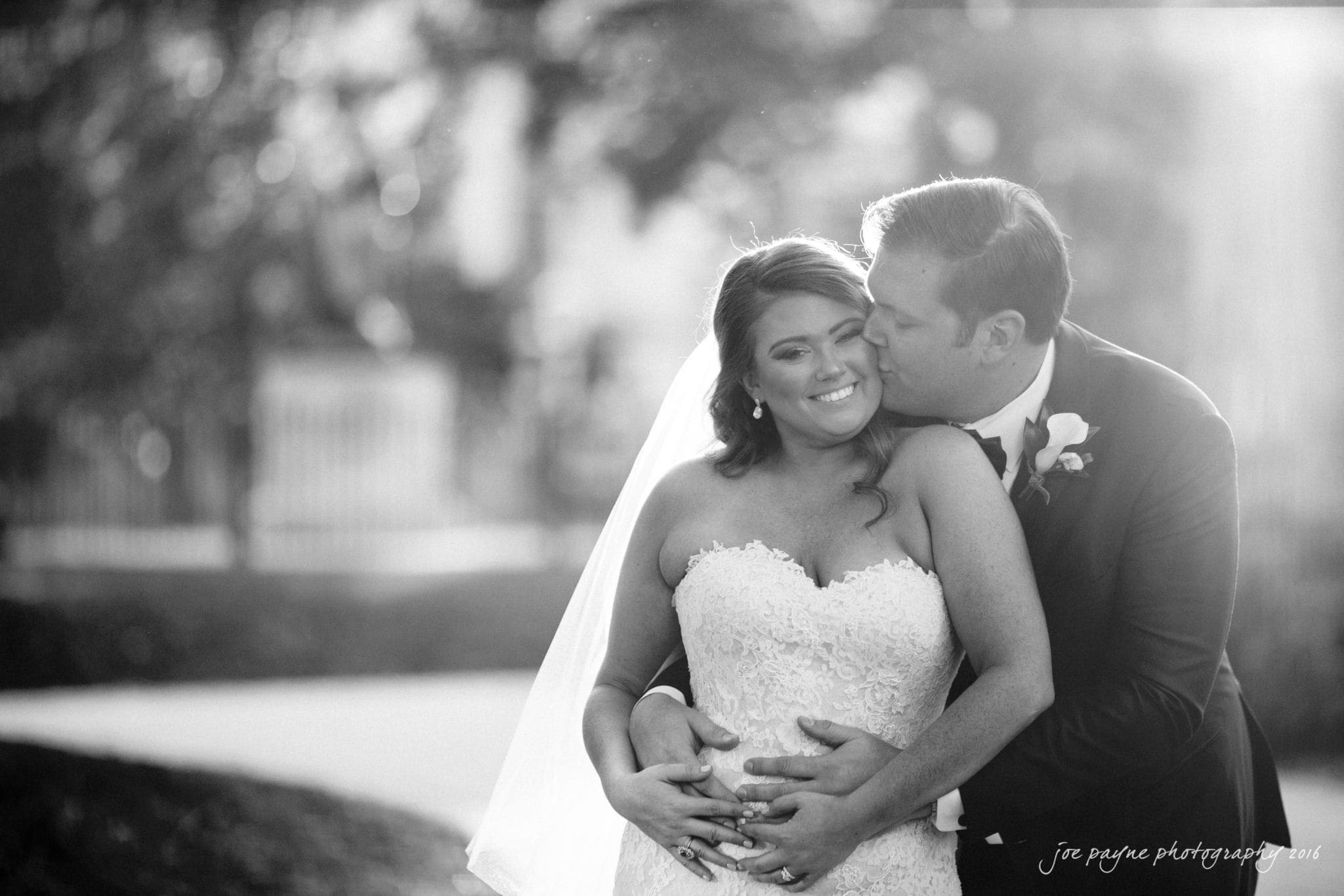 city club wedding photographer - lauren & adam A black-and-white image of a bride and groom. The groom embraces the bride from behind, kissing her cheek. The bride smiles with her eyes closed. Both are dressed in wedding attire, surrounded by blurred outdoor scenery.