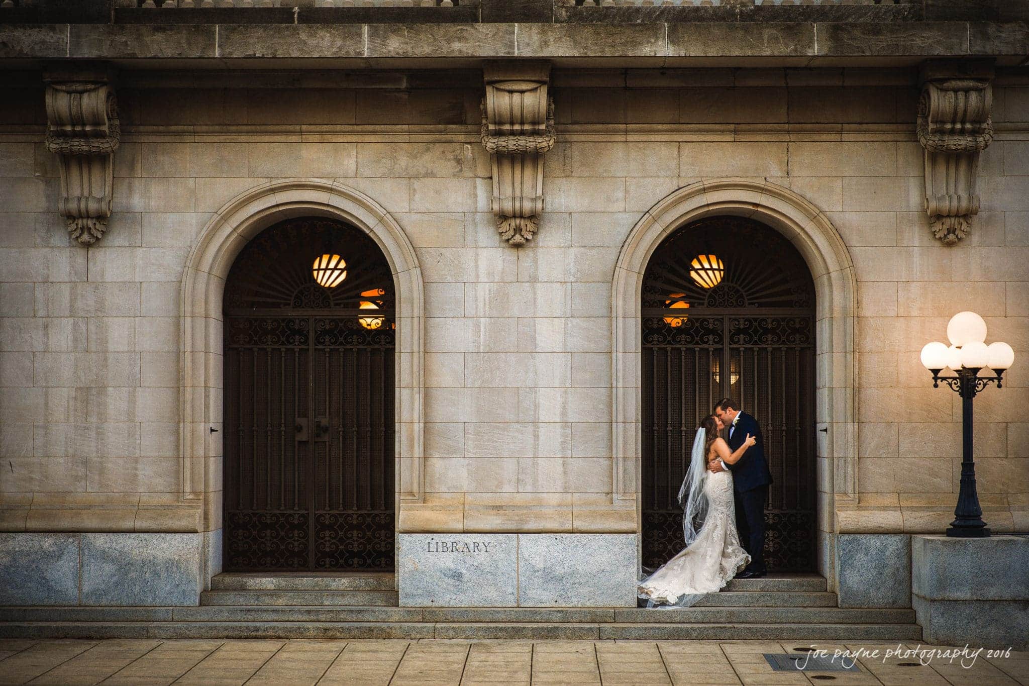 city club wedding photographer - lauren & adam A bride and groom share a kiss in front of a historic library building with arched, gated entrances. The bride wears a long, flowing gown, and the groom is in a dark suit. The scene is warmly lit by nearby street lamps.
