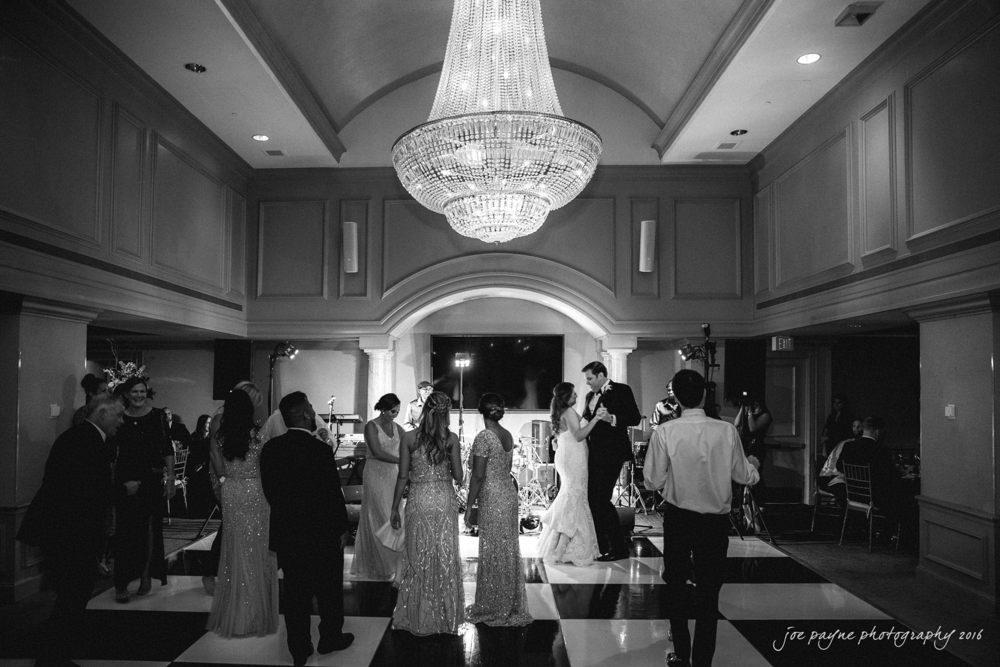 city club wedding photographer - lauren & adam A black and white photo of a ballroom with a large chandelier. Couples, dressed in formal attire, are dancing on a checkered floor. A live band is set up in the background. Photographed by Joe Payne in 2016.