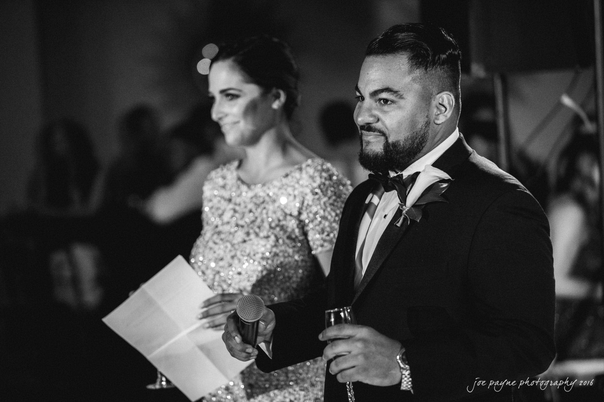 city club wedding photographer - lauren & adam A man in a tuxedo and a woman in a sparkly dress are standing side by side, holding a microphone and a sheet of paper. Both appear to be giving a speech. Its a black and white photo with a lively atmosphere.