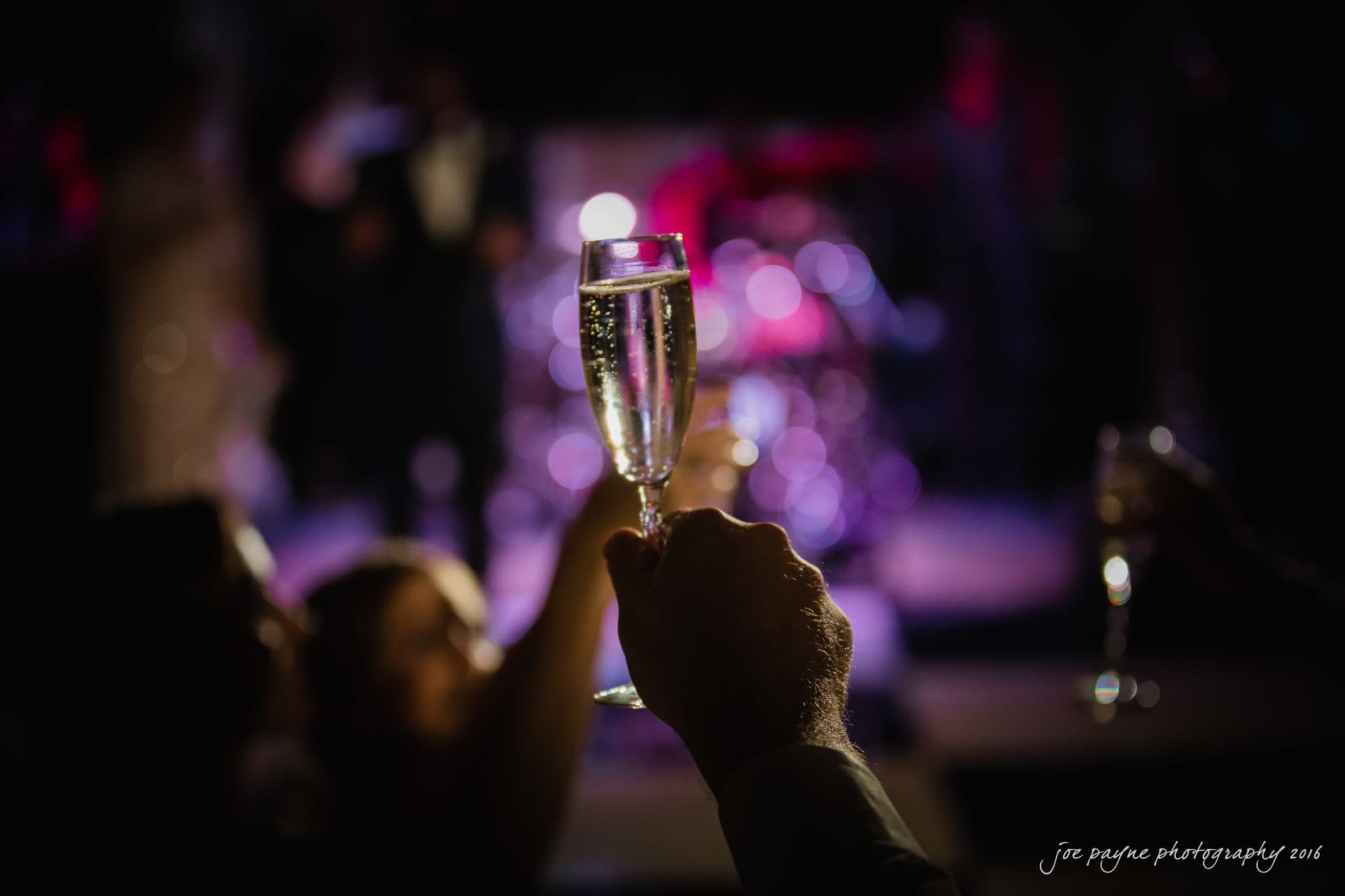 city club wedding photographer - lauren & adam Hands holding up glasses of champagne in a dimly lit setting with blurred figures and colorful lights in the background, creating a festive atmosphere.